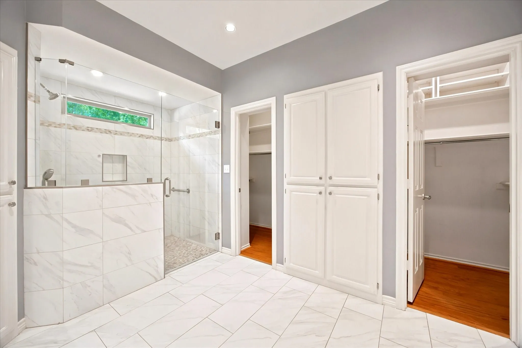 Spacious bath featuring a glass-enclosed shower with a tiled bench and pebble floor, white tile flooring, recessed lighting, built-in cabinetry, and two walk-in closets with wood-finish flooring