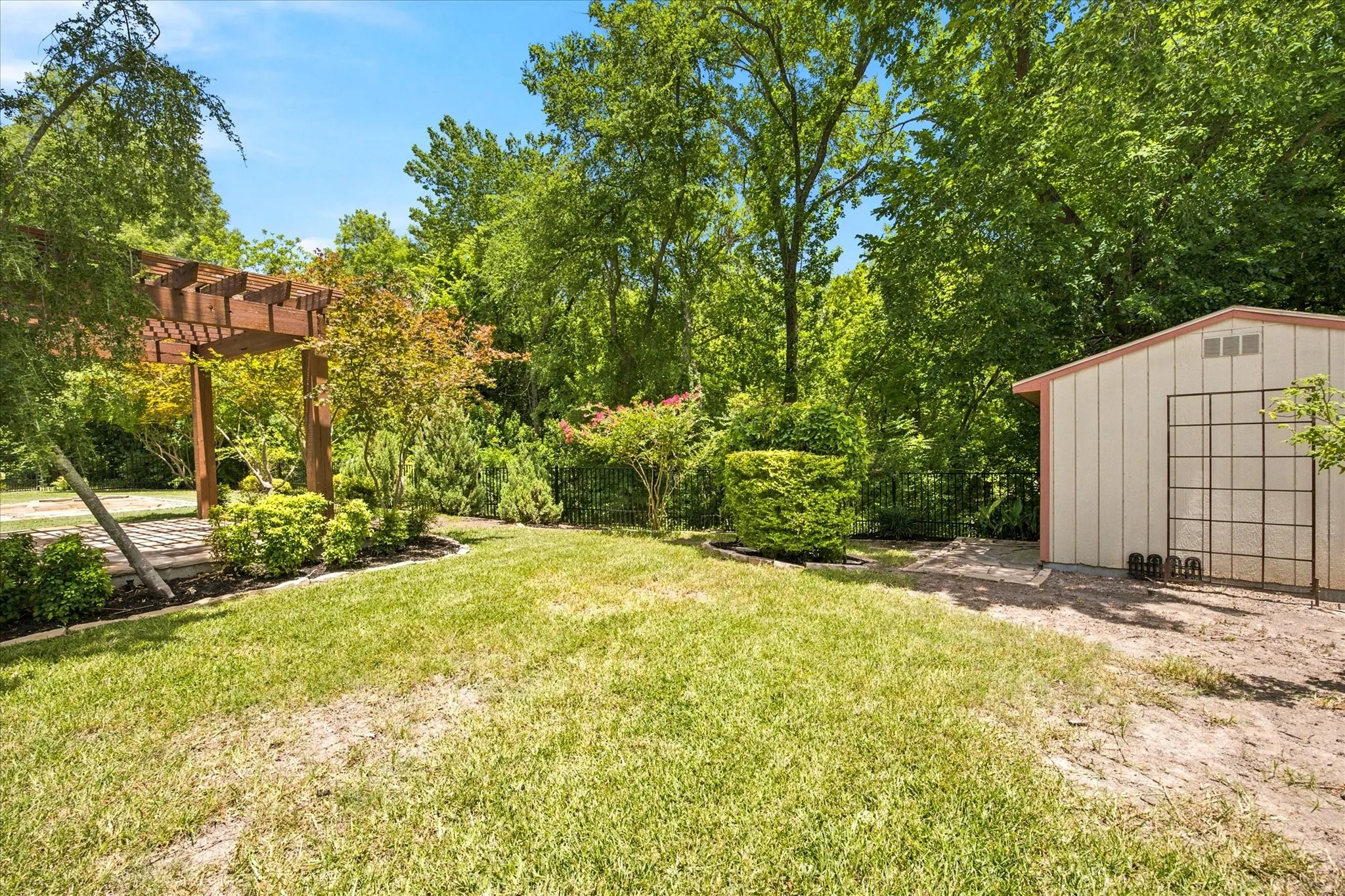 Expansive backyard featuring a wood pergola with a stone paver base, mature trees, a manicured lawn, and a storage shed with vertical siding