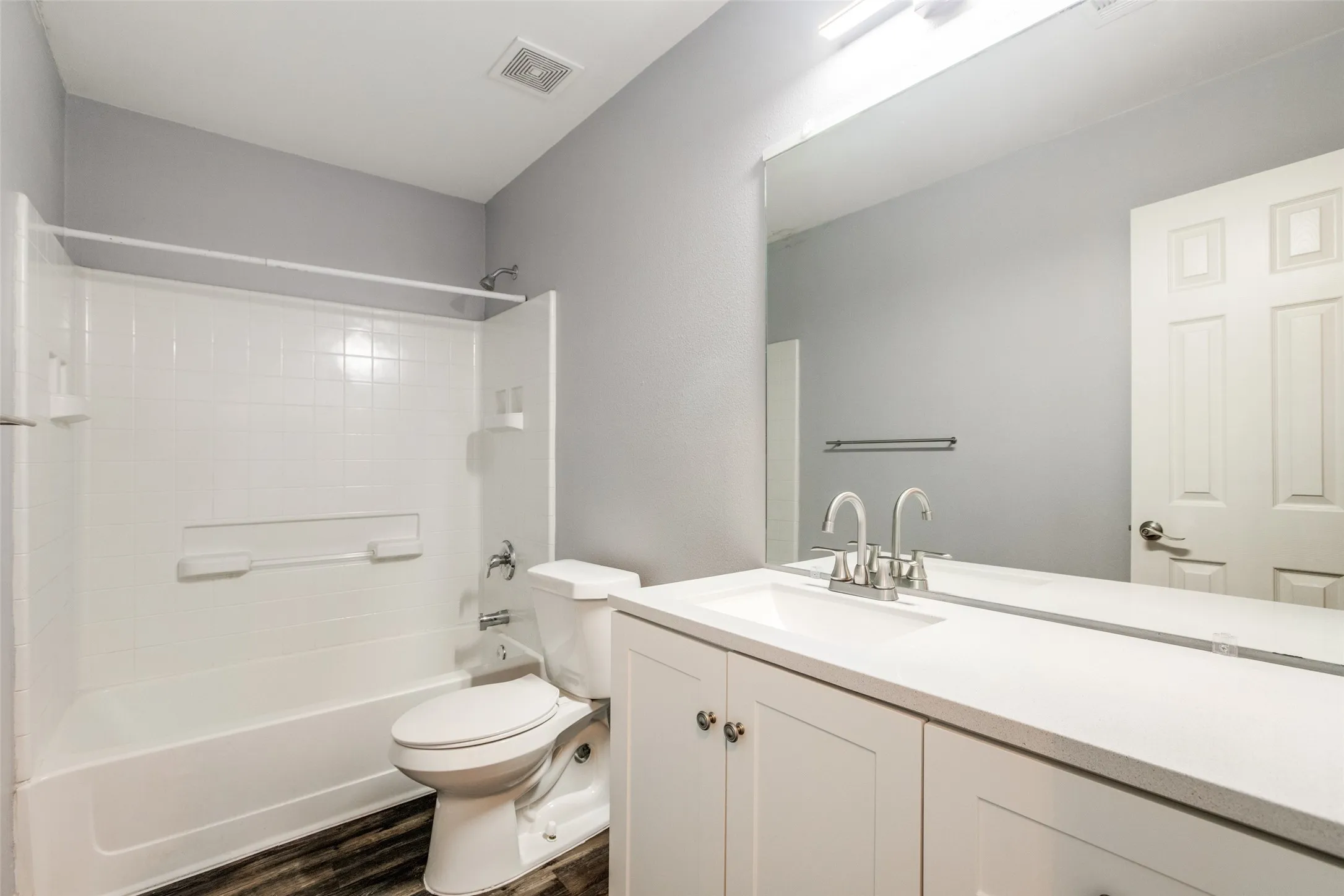Bathroom featuring a white vanity with integrated sink, a full-width mirror, a white bathtub with integrated shower, and wood-finish flooring