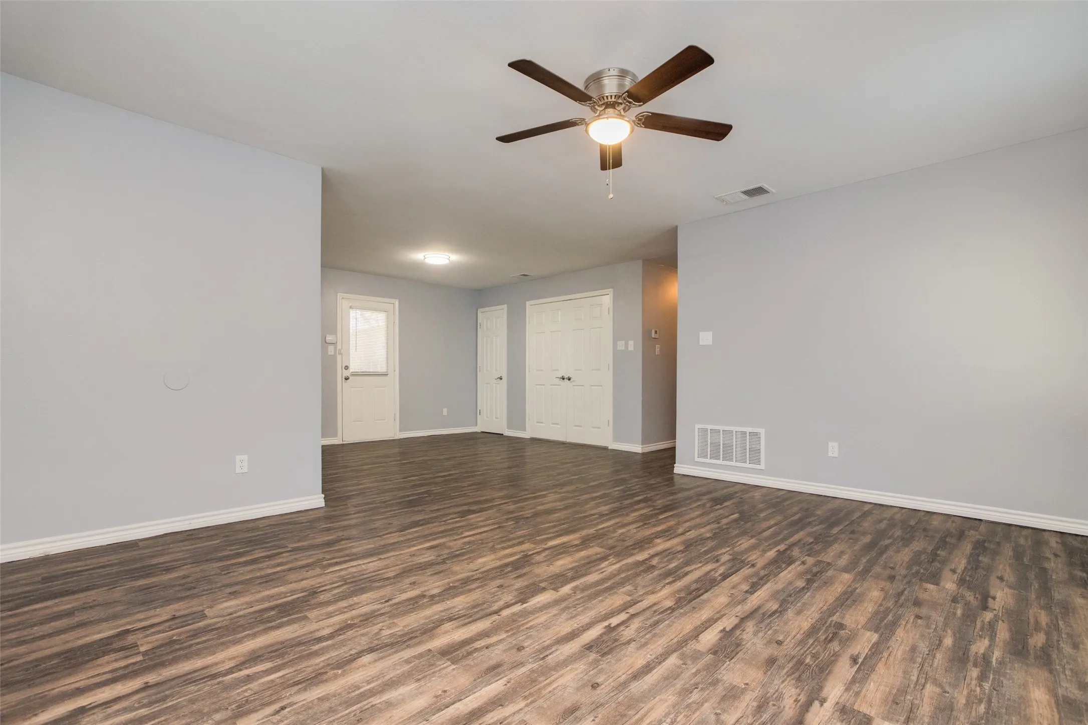 Spacious room featuring wood-finish flooring, light grey walls, white baseboards, a ceiling fan with integrated lighting, and multiple white doors