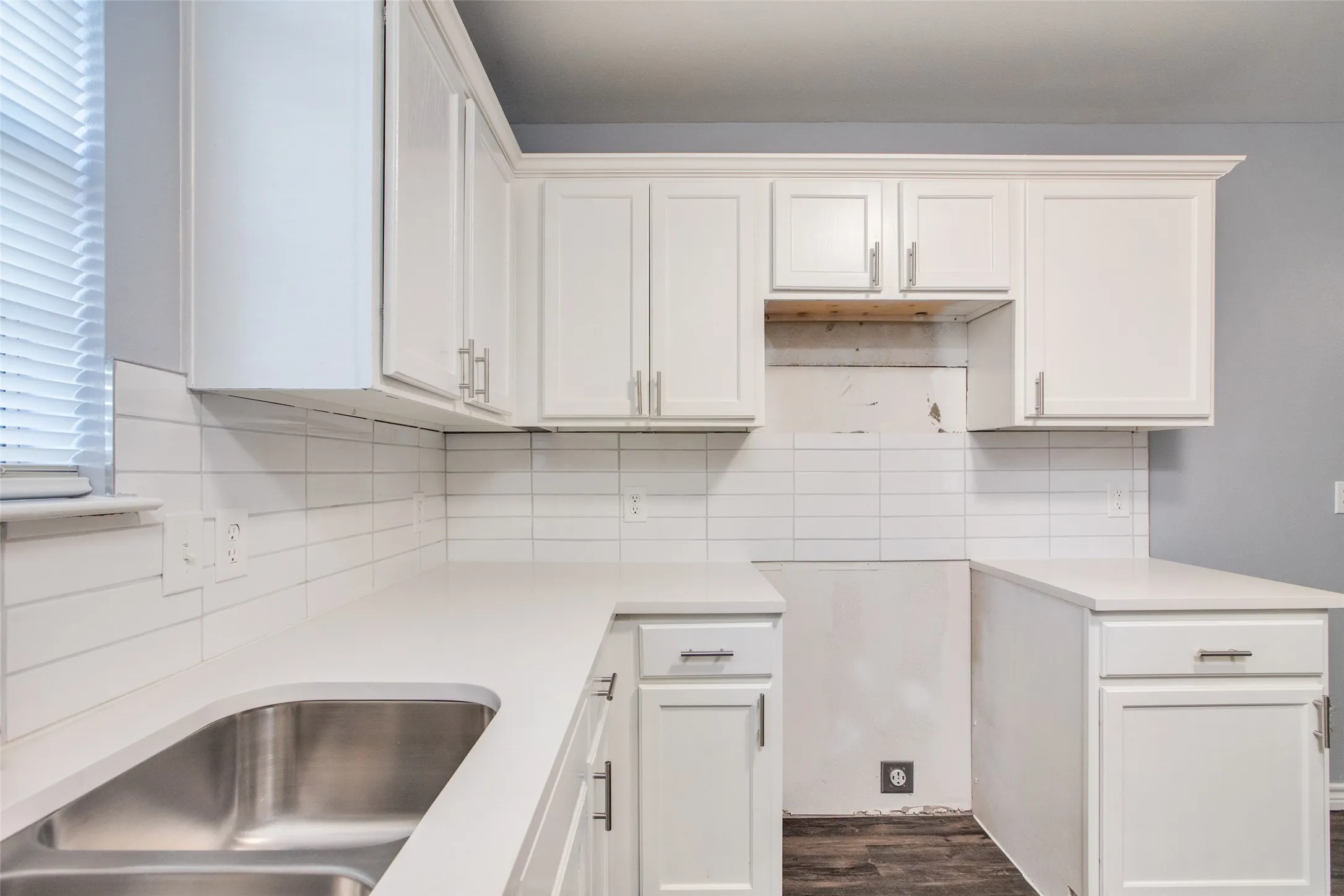 Kitchen featuring white cabinetry, light-toned countertops, a stainless steel double-basin sink, white subway tile backsplash, and wood-finish flooring