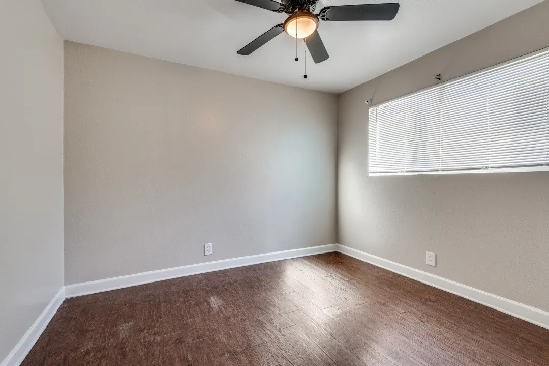 Spacious room featuring wood-finish flooring, light grey painted walls, white baseboards, and a ceiling fan with integrated lighting