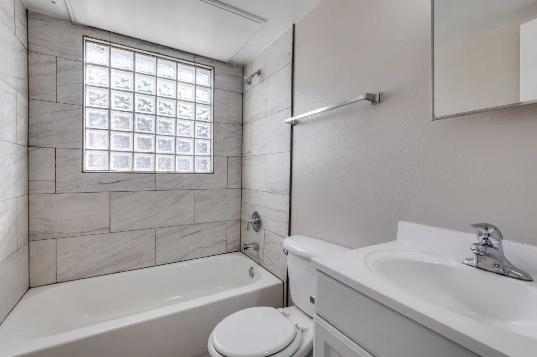 Bathroom featuring a tub/shower combination, glass block window, chrome fixtures, white vanity with integrated sink, and large format tile surround