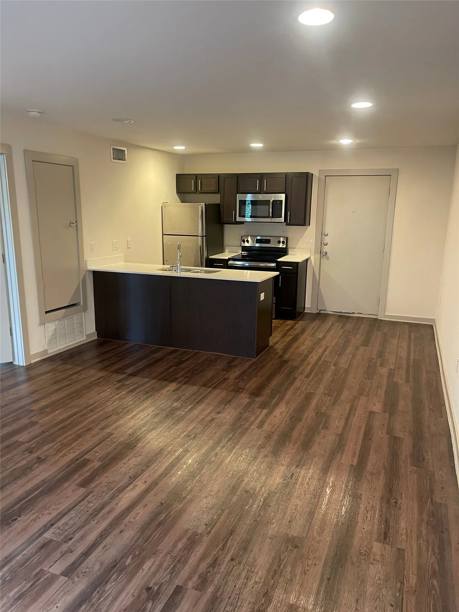 Open-concept living space featuring wood-finish flooring, a kitchen island with integrated sink, dark wood cabinetry, stainless steel appliances, and recessed ceiling lighting