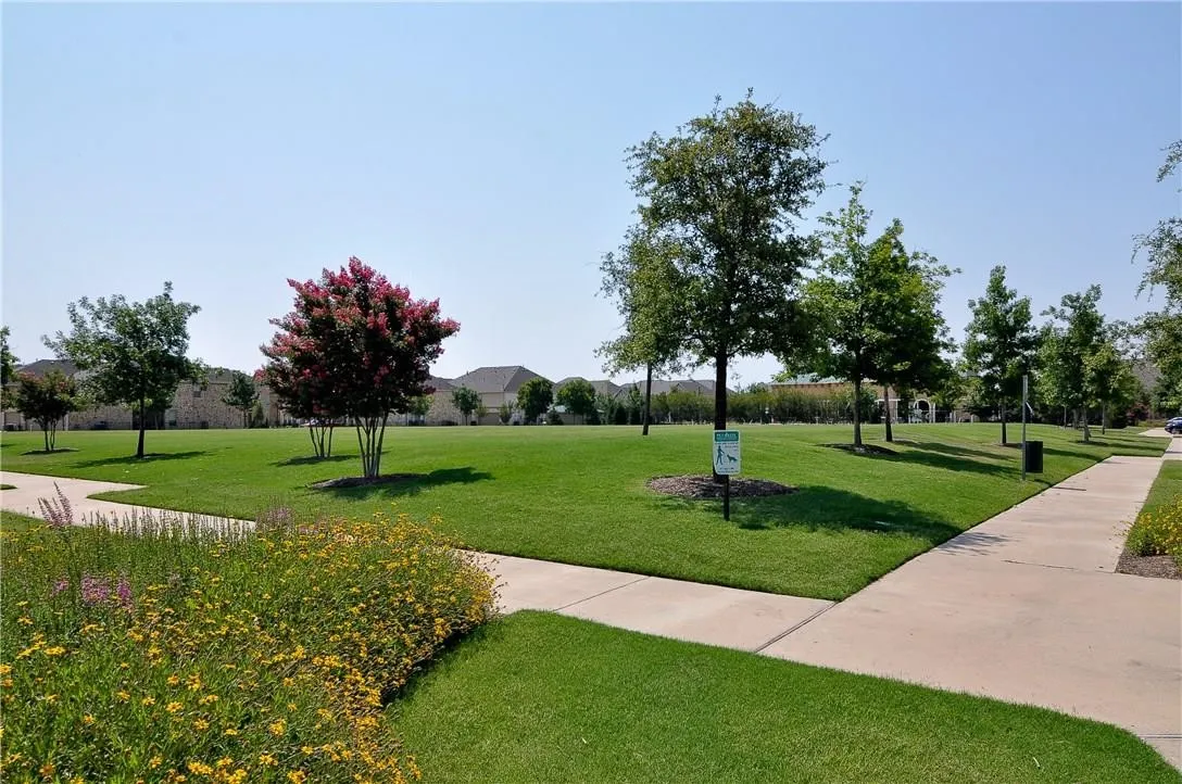 Expansive green space featuring mature trees, a flowering tree with pink blossoms, concrete pathways, and a landscaped border with mixed flora