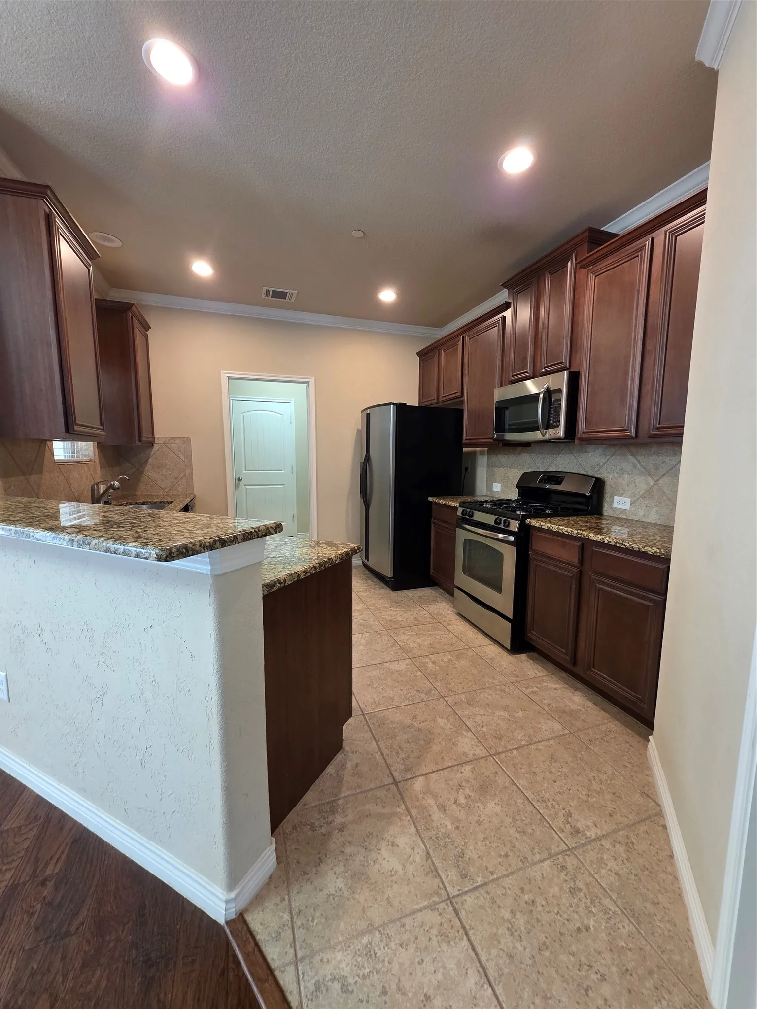 Modern kitchen featuring rich wood cabinetry, stainless steel appliances, granite countertops, recessed ceiling lighting, and tile flooring throughout the open layout with a centered island workspace