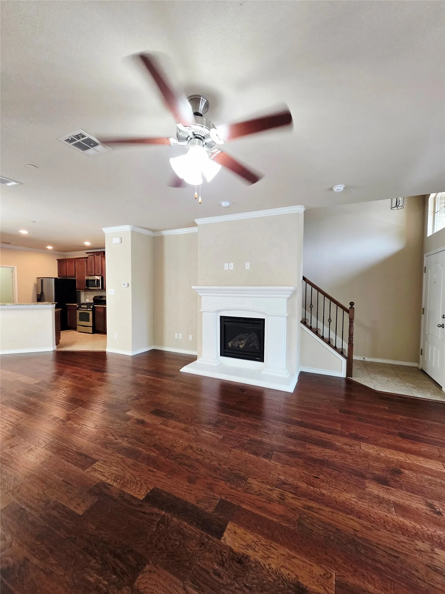 Wood-finish flooring throughout, featuring a fireplace with a white mantel, an overhead ceiling fan, and a wood and wrought iron staircase