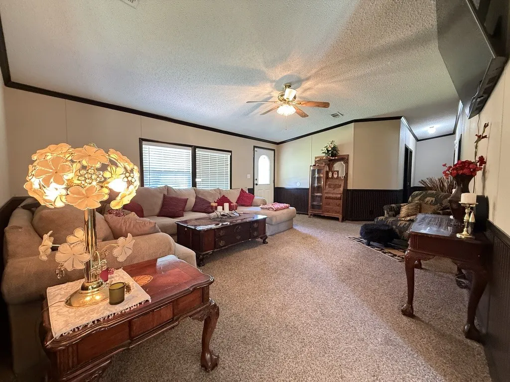 Spacious living area featuring neutral carpet flooring, a ceiling fan, and prominent window with blinds