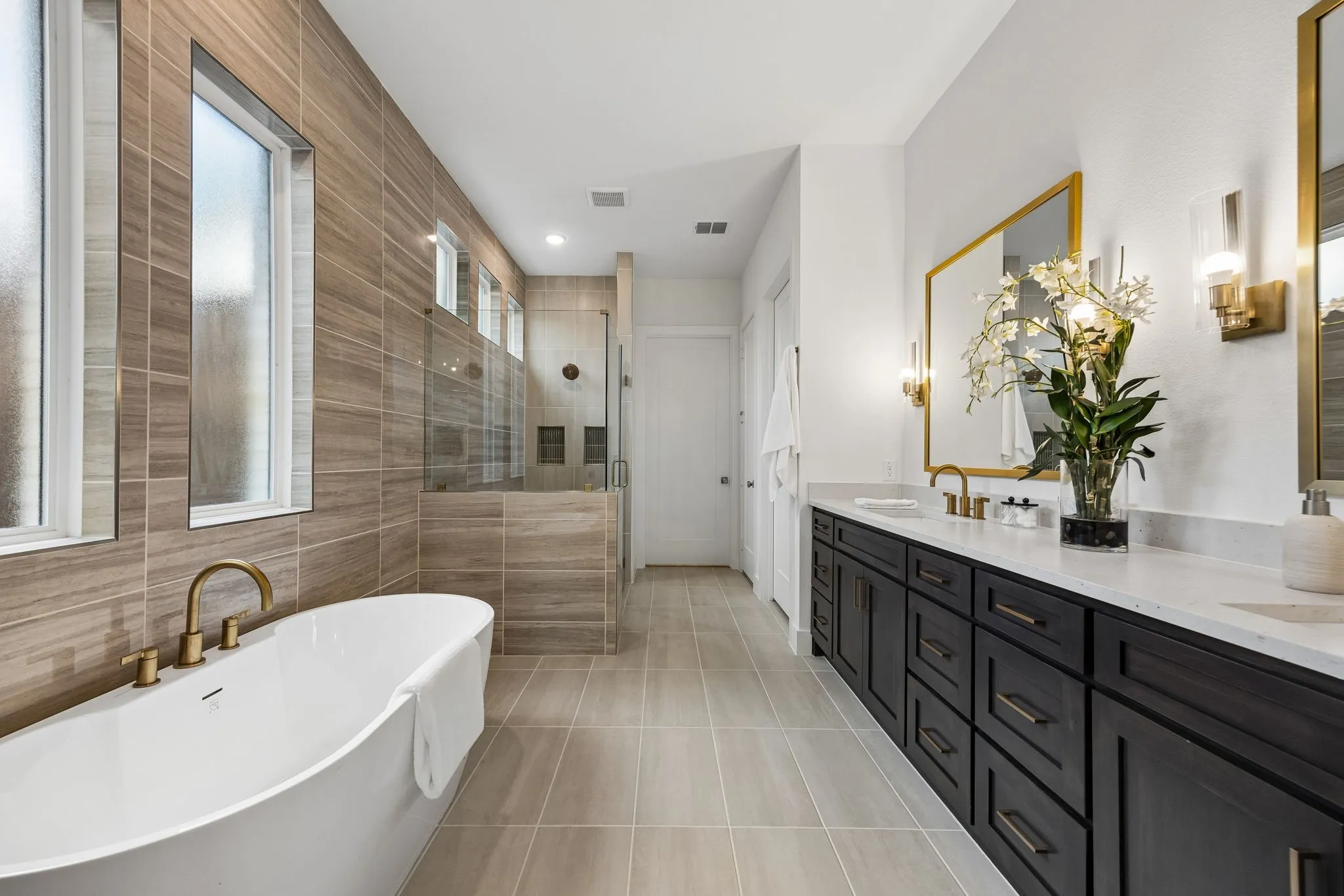 Freestanding soaking tub with brass fixtures, expansive tile-clad shower with glass enclosure, dual vanity with dark wood-finish cabinetry, light stone-finish countertop, and gold-framed mirrors