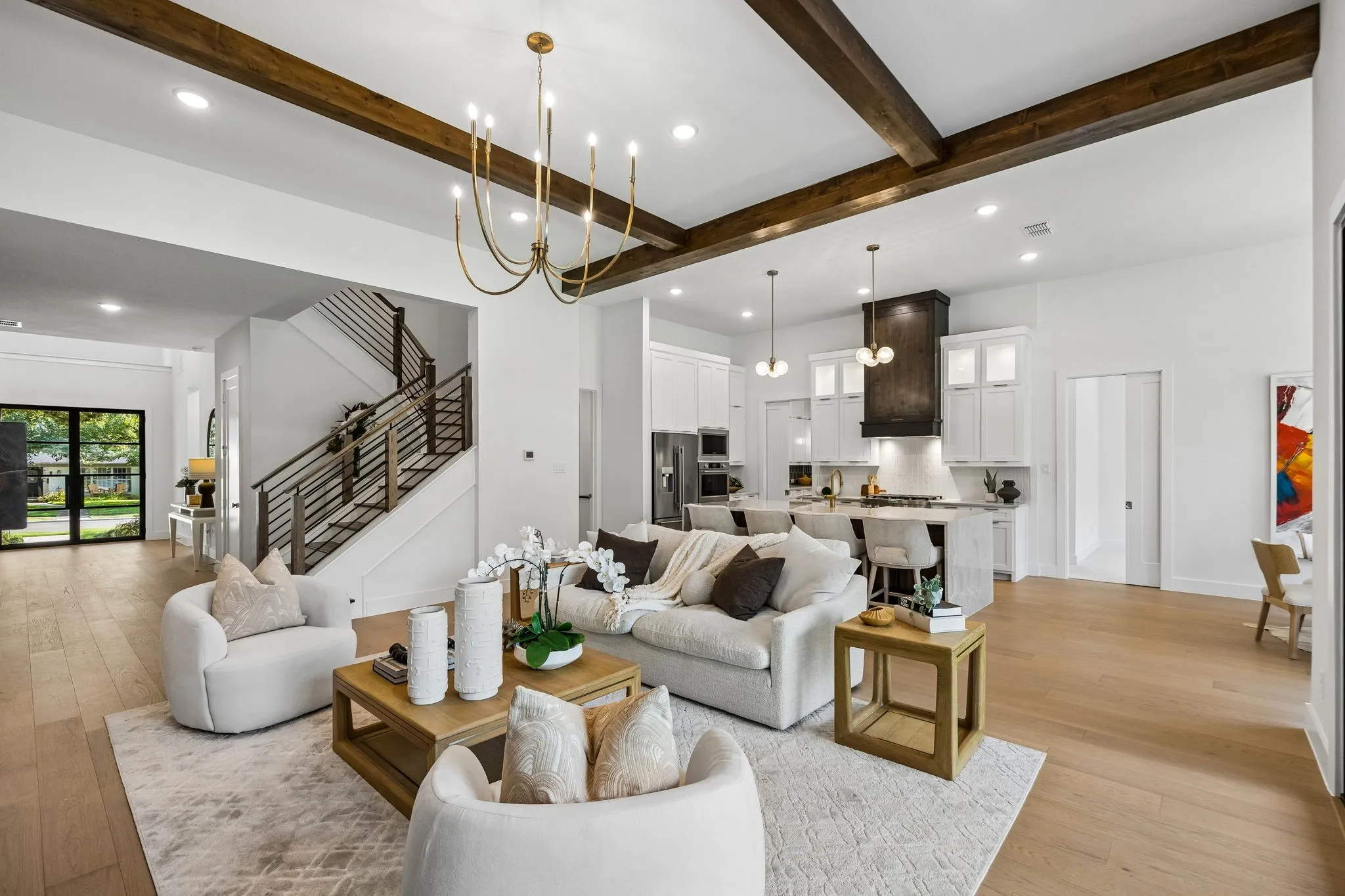 Open-concept living space with wood-finish flooring, exposed ceiling beams, a brass chandelier, a modern staircase with metal railings, and a kitchen featuring white cabinetry and a dark wood range hood