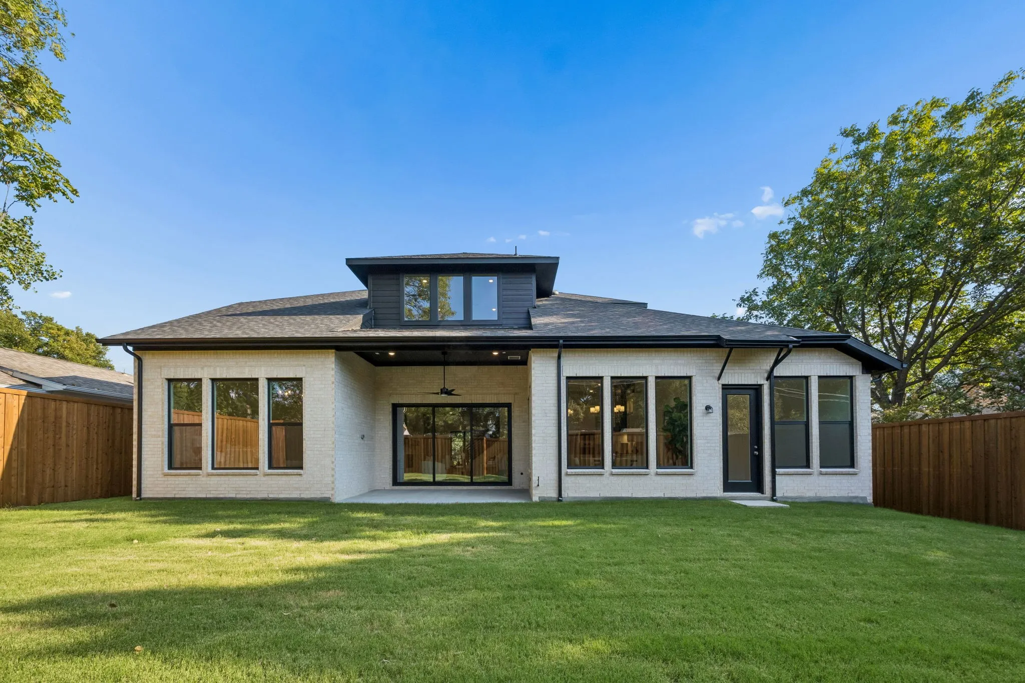 Expansive backyard featuring a manicured lawn, brick exterior, multiple large windows, and a covered patio with a ceiling fan