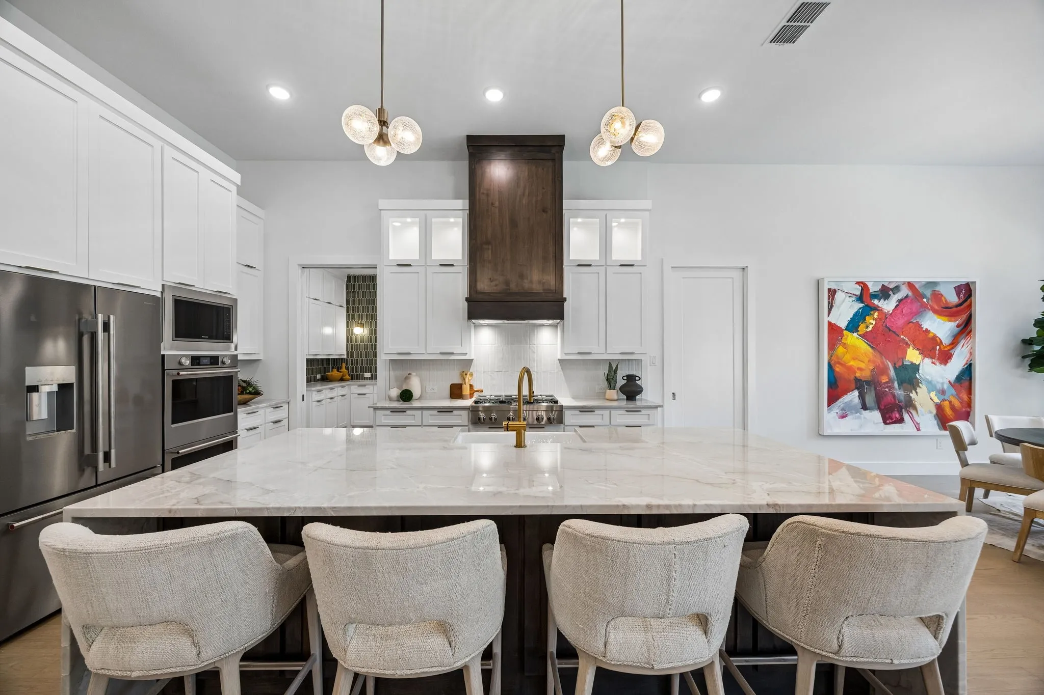 Expansive kitchen island with a stone countertop, white shaker cabinetry, stainless steel appliances, a wood-finish range hood, and contemporary pendant lighting