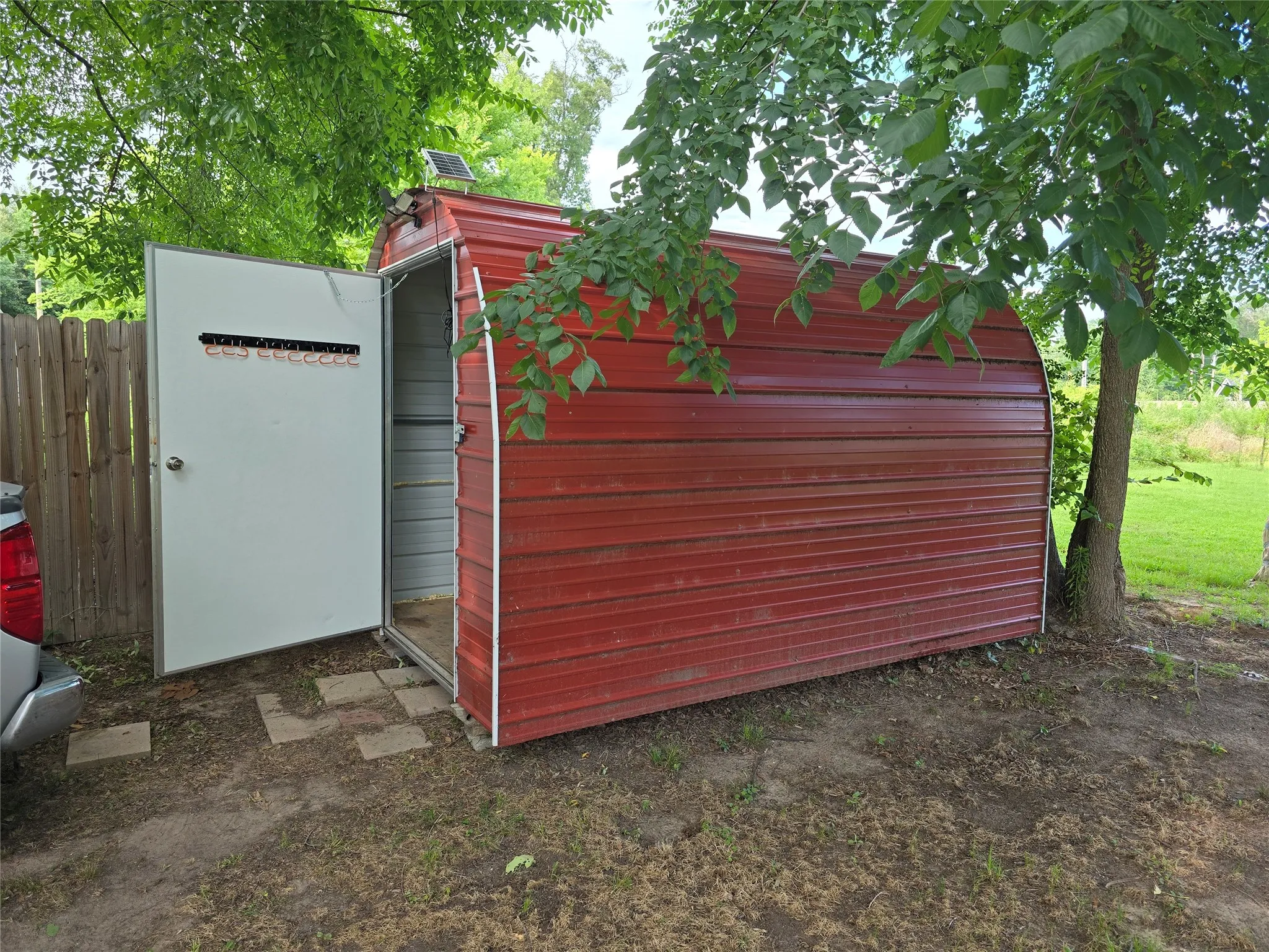 Red corrugated metal storage shed with a white entry door, solar panel, and a wooden privacy fence