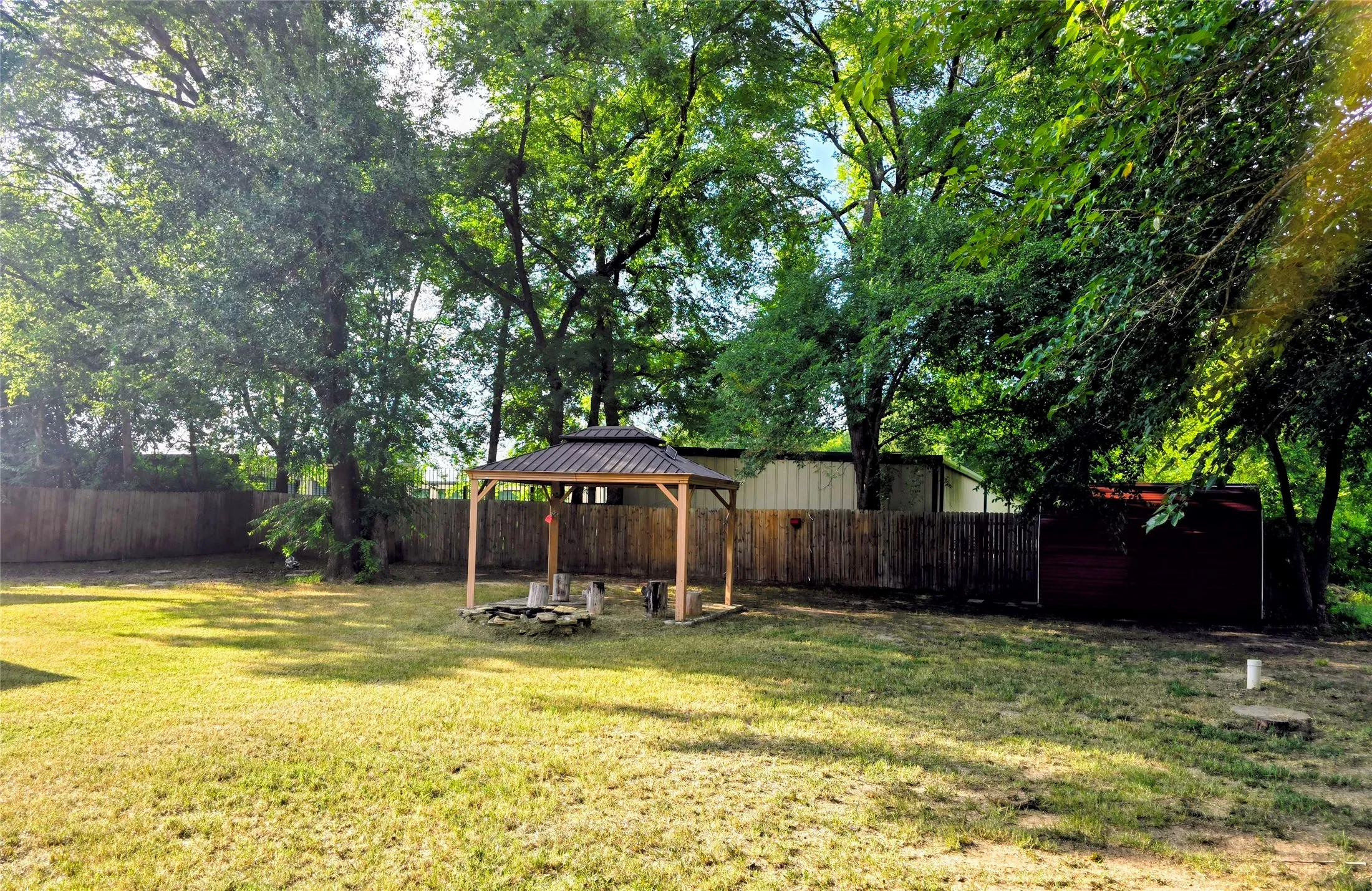 Expansive backyard featuring a central gazebo with a metal roof, a stone-lined fire pit, and mature trees providing natural shading