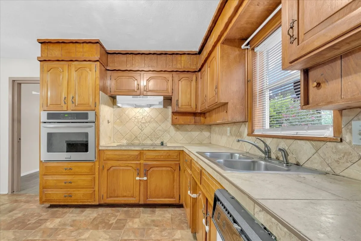 Kitchen featuring wood cabinetry, stainless steel wall oven, tile countertops, tile backsplash, and a double basin stainless steel sink