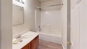 Bathroom featuring a wood-finish vanity with an integrated sink, a wall-mounted mirror, overhead lighting, a shower/tub combination, and wood-finish flooring