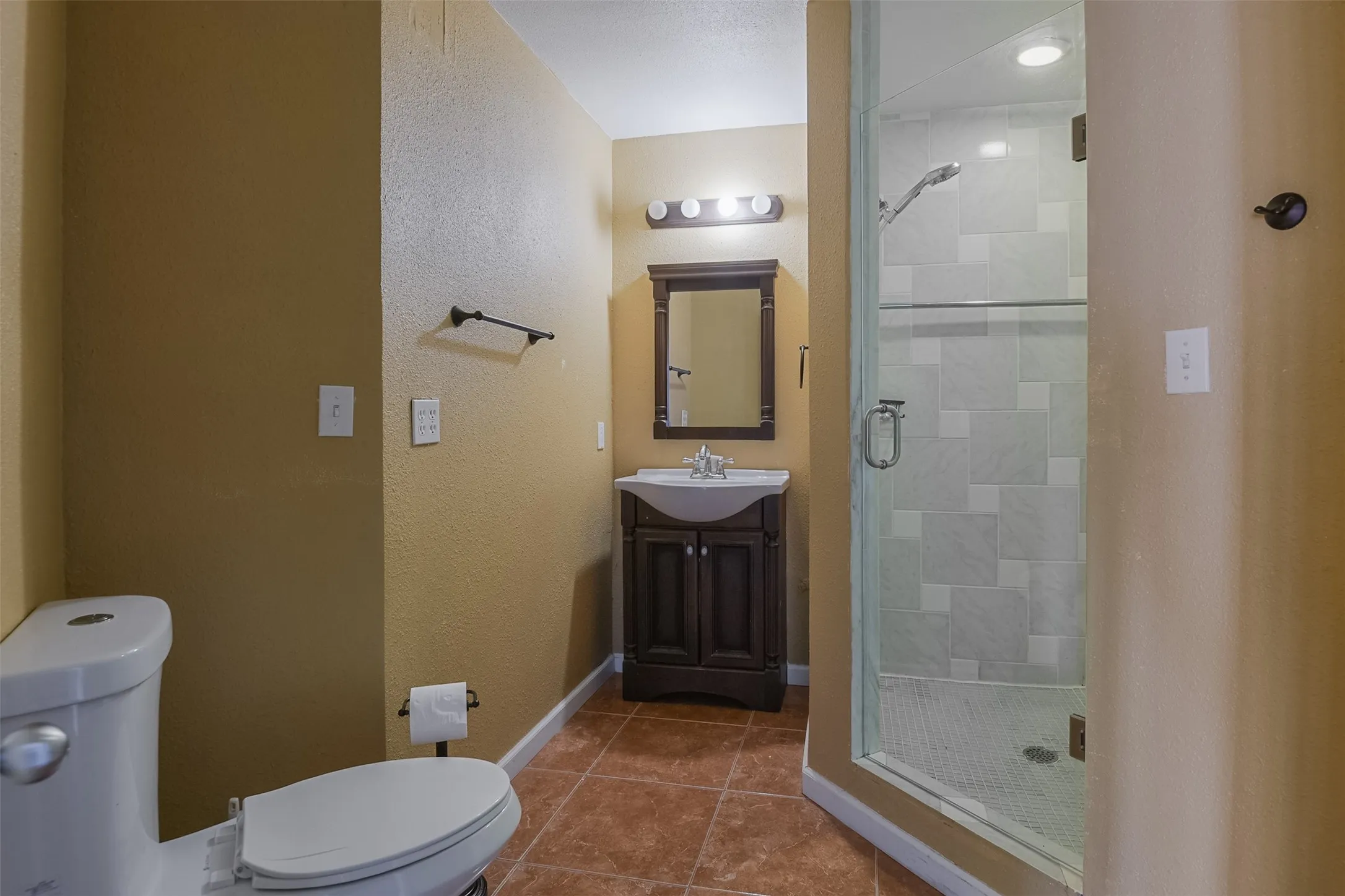Spa-inspired primary bathroom featuring a frameless glass shower enclosure with tile surround, dark wood vanity with integrated sink and countertop, dual vanity lighting, ceramic tile flooring, and a separate water closet for added convenience
