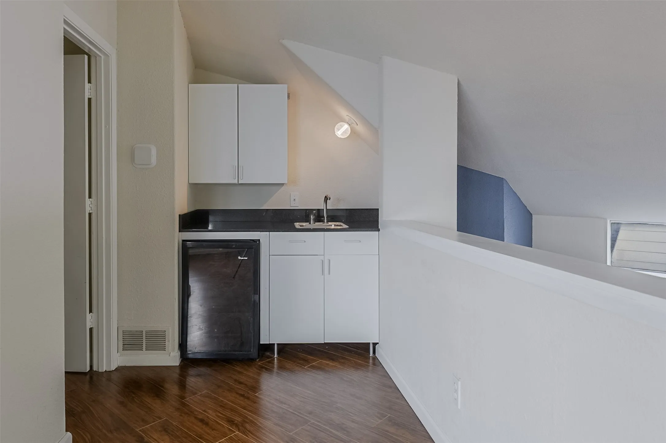 Compact wet bar featuring a single-basin sink, dark countertop, white cabinetry, and wood-finish flooring