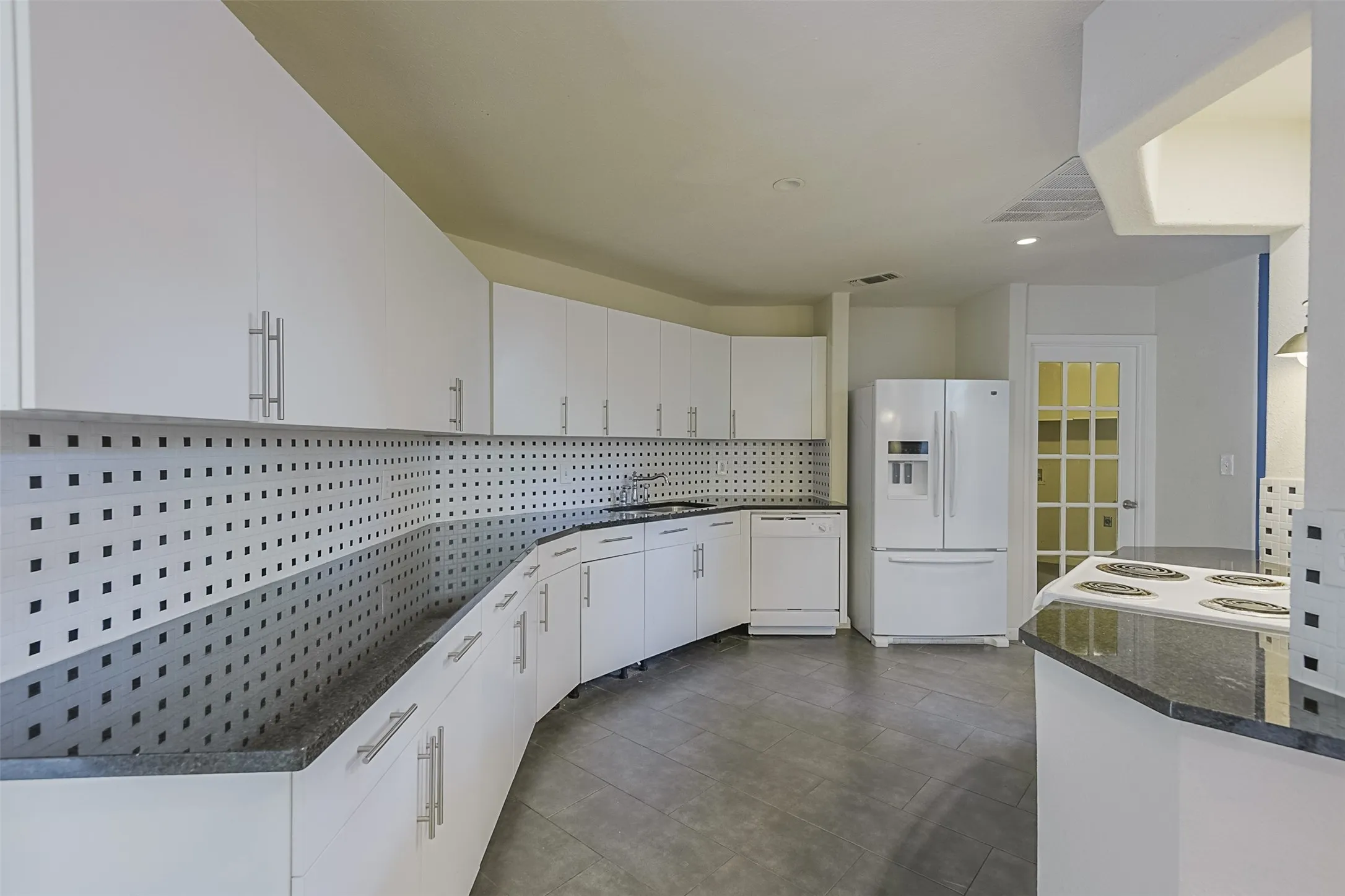 Modern kitchen featuring white cabinetry with polished hardware, perforated stainless steel backsplash, concrete flooring, curved countertop design, and stainless steel appliances including a refrigerator and range