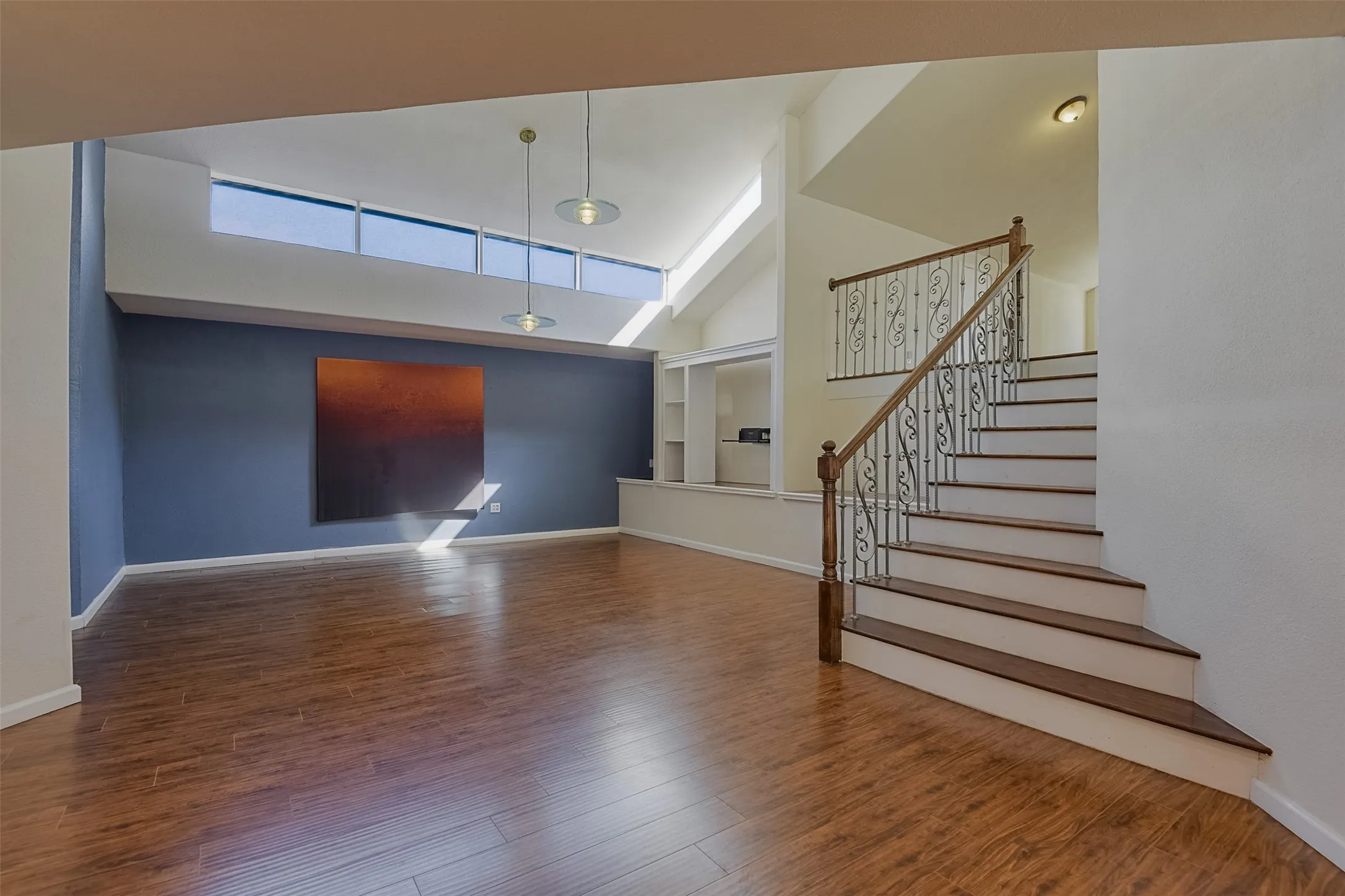 Wood-finish flooring throughout, multi-level ceiling, built-in shelving, staircase with decorative wrought iron railing, and high transom windows