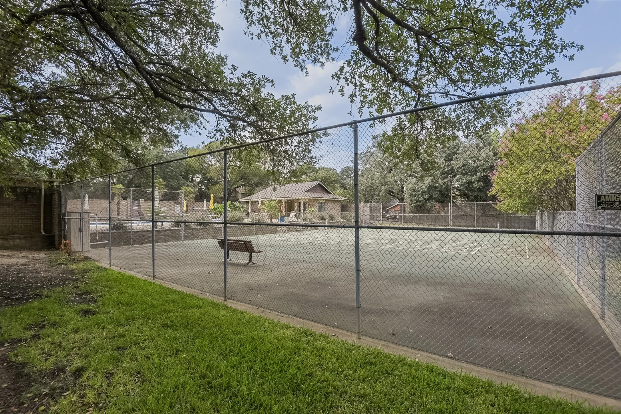 Fenced tennis court featuring a hard surface, surrounding mature trees, and a distant pool area