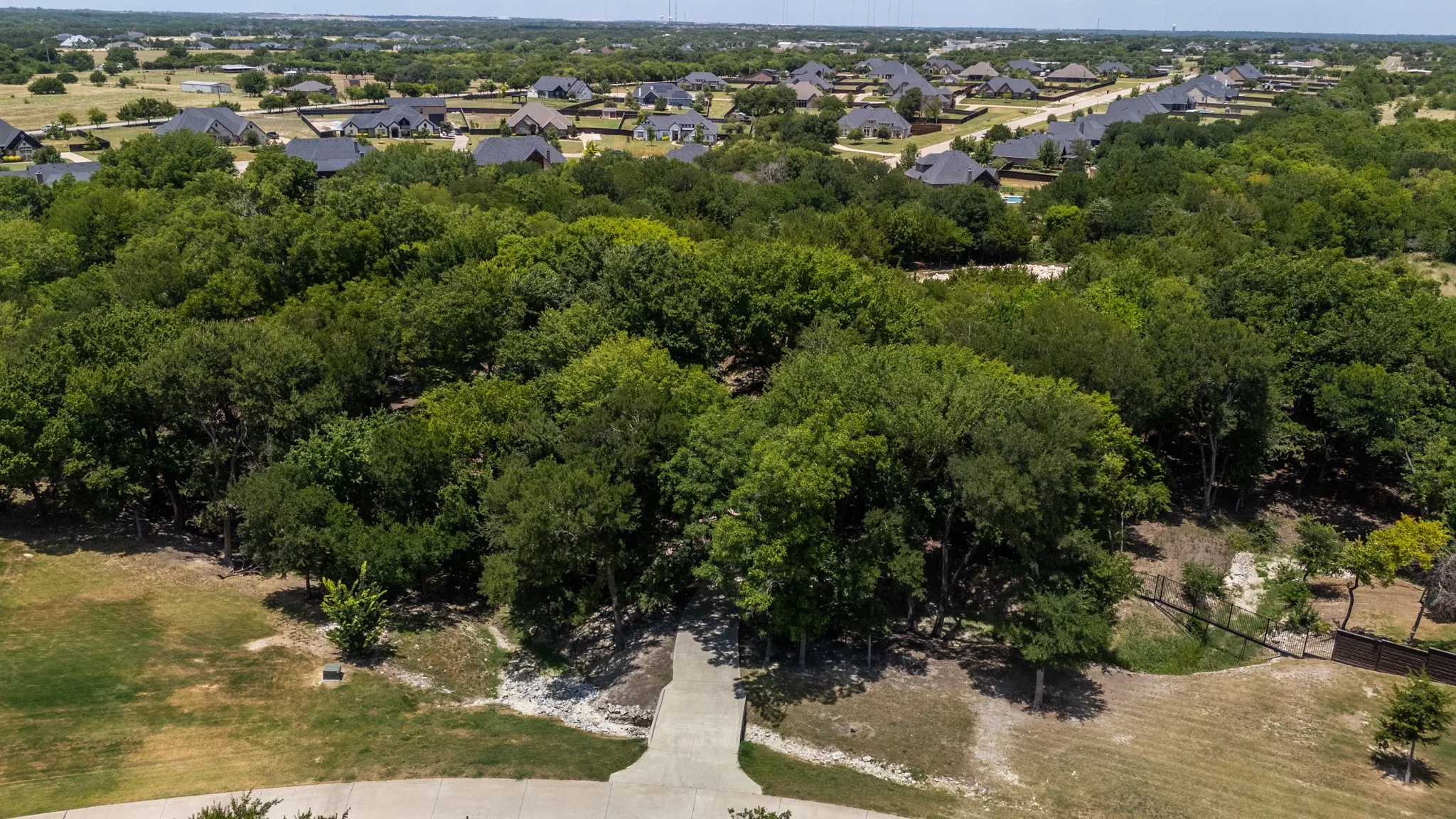 Expansive treed lot featuring a concrete driveway, surrounding green spaces, and a privacy fence