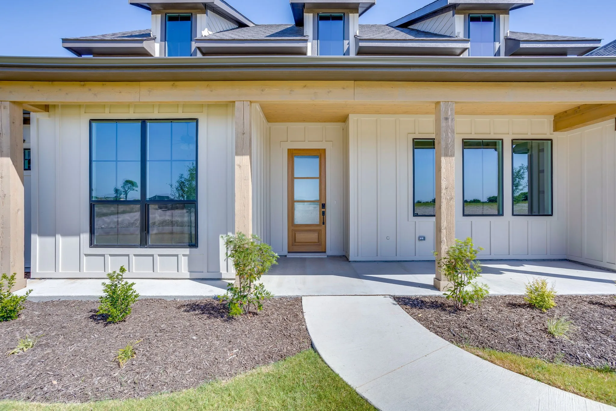 Exterior featuring a light wood front door with vertical glass panels, board-and-batten siding, black-framed windows, and exposed wood porch columns