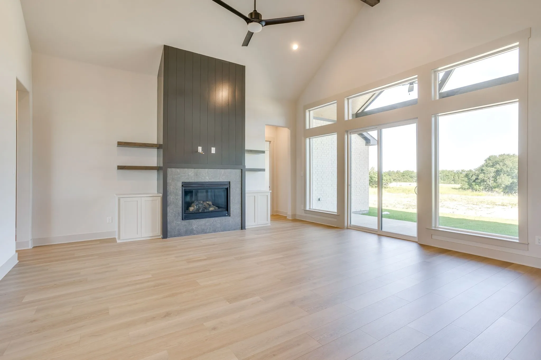 Vaulted ceiling living space featuring a dark gray shiplap fireplace surround, built-in shelving, wood-finish flooring, and expansive windows with a sliding glass door