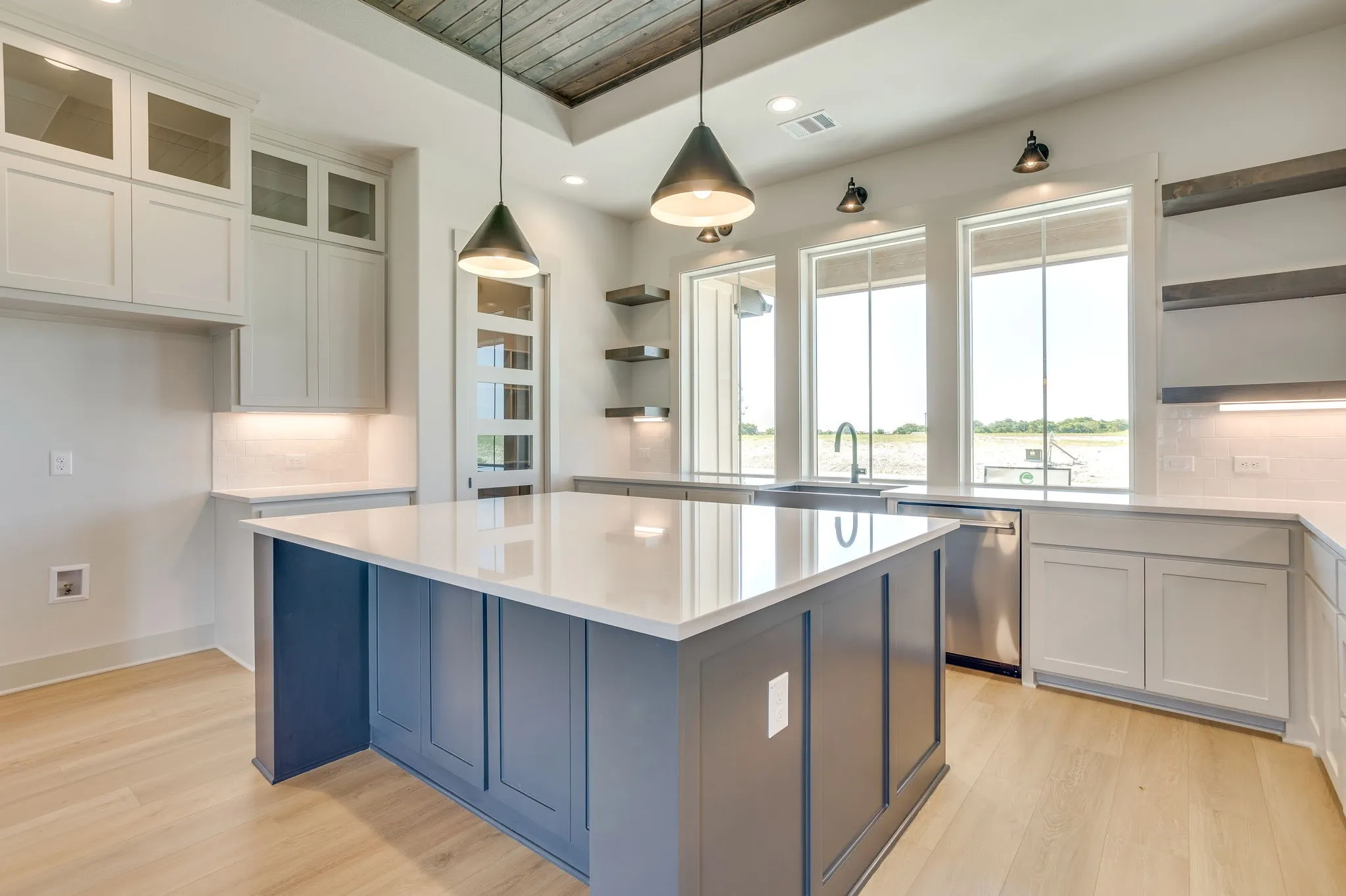 Spacious kitchen featuring a large central island with a light-toned countertop, light wood-finish flooring, white cabinetry, integrated stainless steel appliances, and abundant natural light from multiple windows