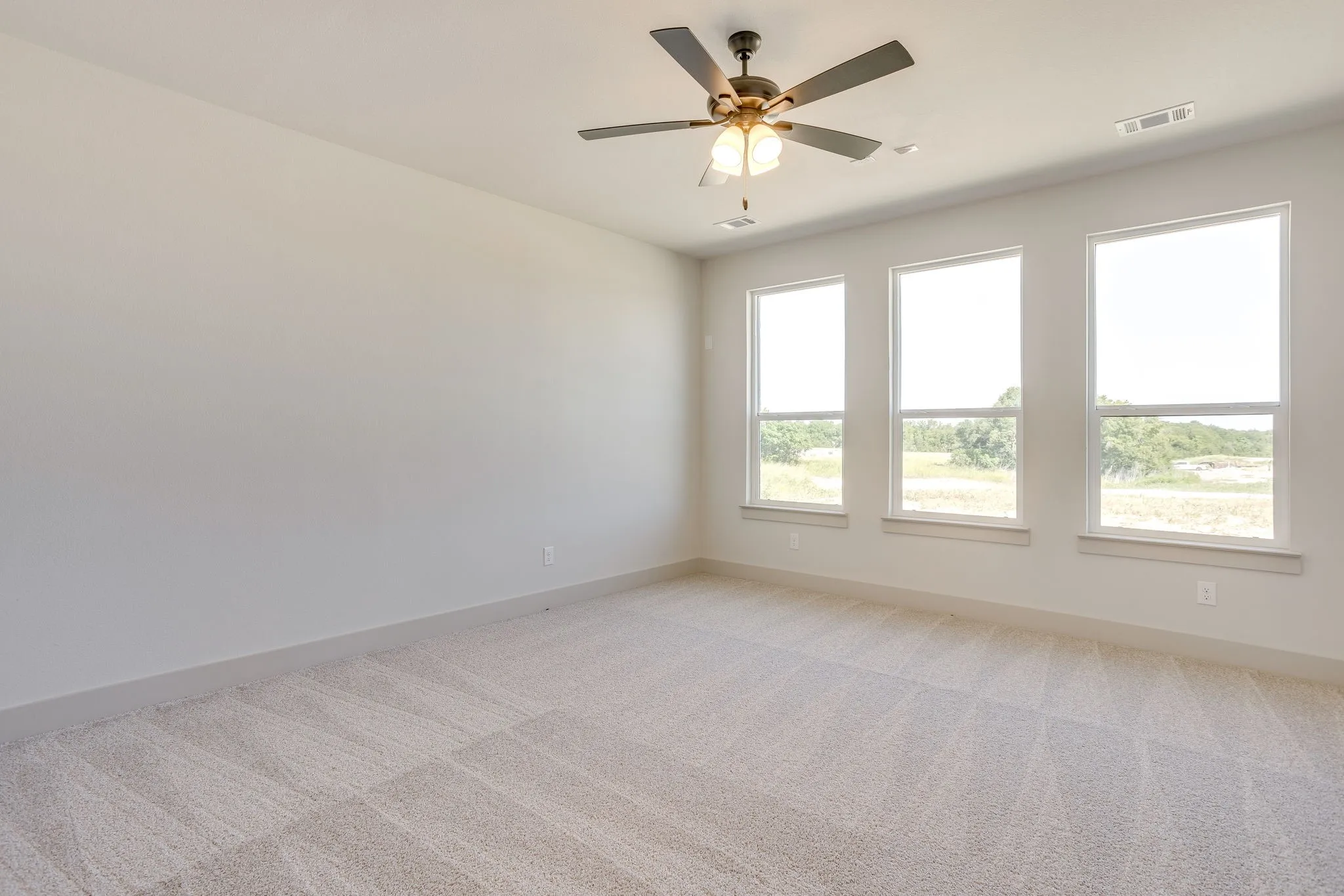 Spacious room featuring three large windows, neutral-toned carpeting, light gray walls, and a ceiling fan with integrated lighting