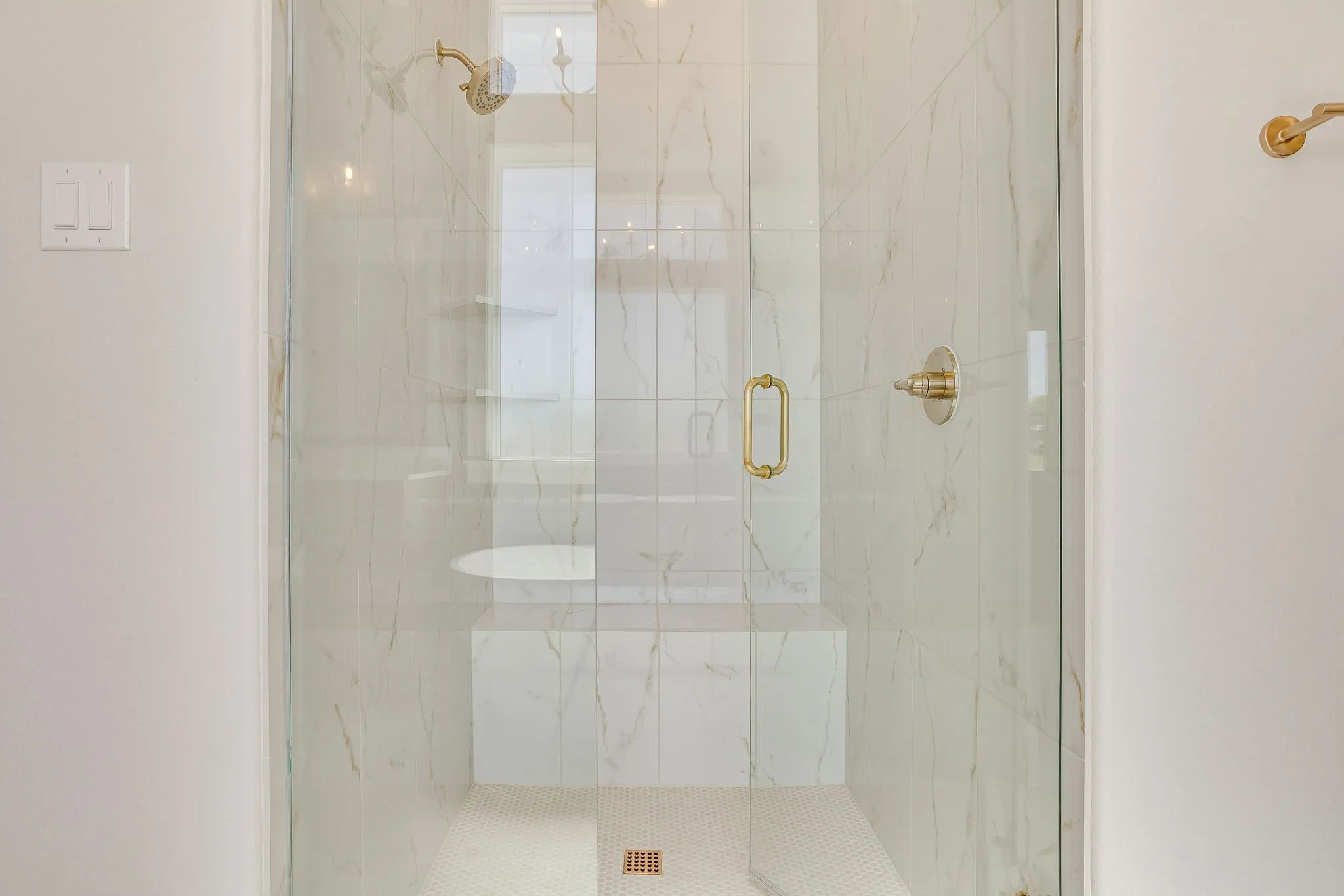Frameless glass shower enclosure featuring brass fixtures, marble-look tile walls, a built-in bench, and a mosaic tile floor
