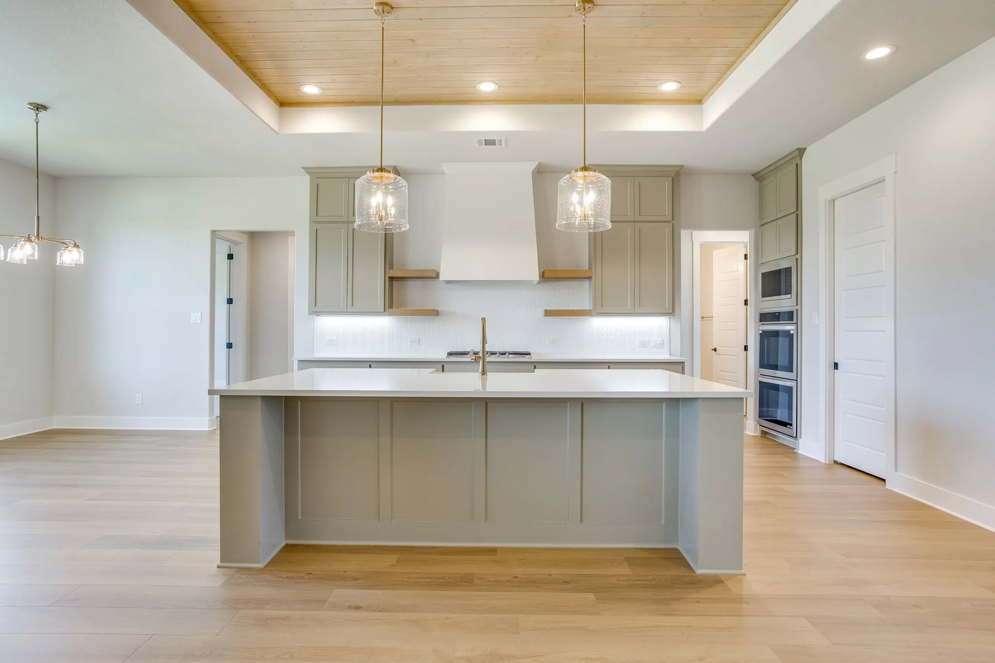 Kitchen featuring a large central island with a light-toned countertop, recessed lighting, wood-finish flooring, a wood-paneled ceiling accent, and stainless steel built-in ovens