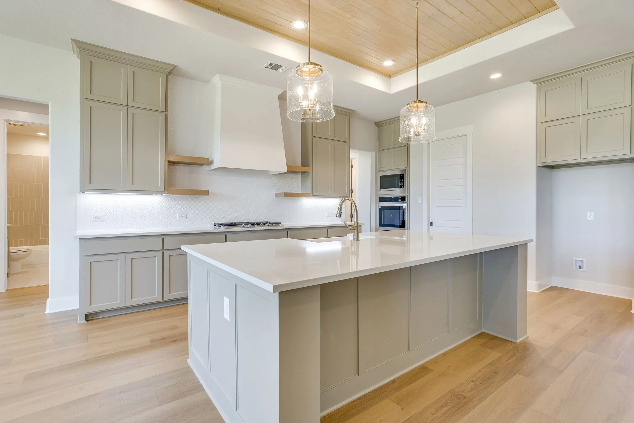 Spacious kitchen with a central island featuring a sink, light wood-finish flooring, light gray cabinetry, built-in stainless steel appliances, and a wood-panelled ceiling with recessed lighting and pendant fixtures