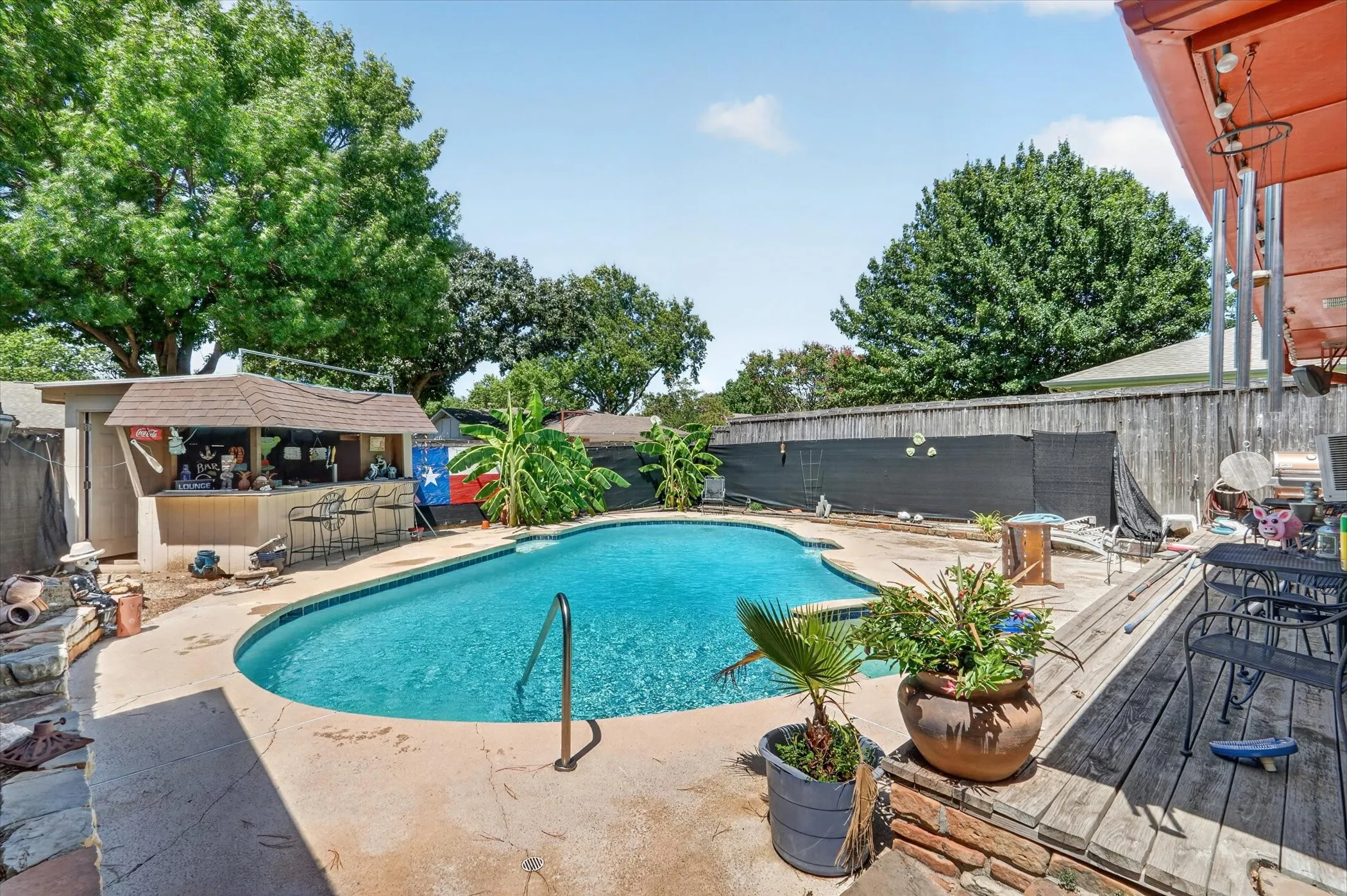 Backyard oasis featuring an in-ground swimming pool with integrated steps, a dedicated outdoor bar structure with a shingled roof, extensive concrete patio surround, a raised wood deck, and mature perimeter trees
