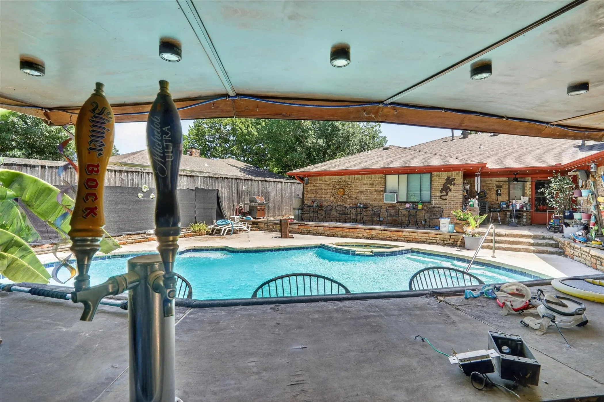 Outdoor covered patio with built-in bar, integrated beverage taps, and recessed lighting overlooking a fenced swimming pool with a stone surround