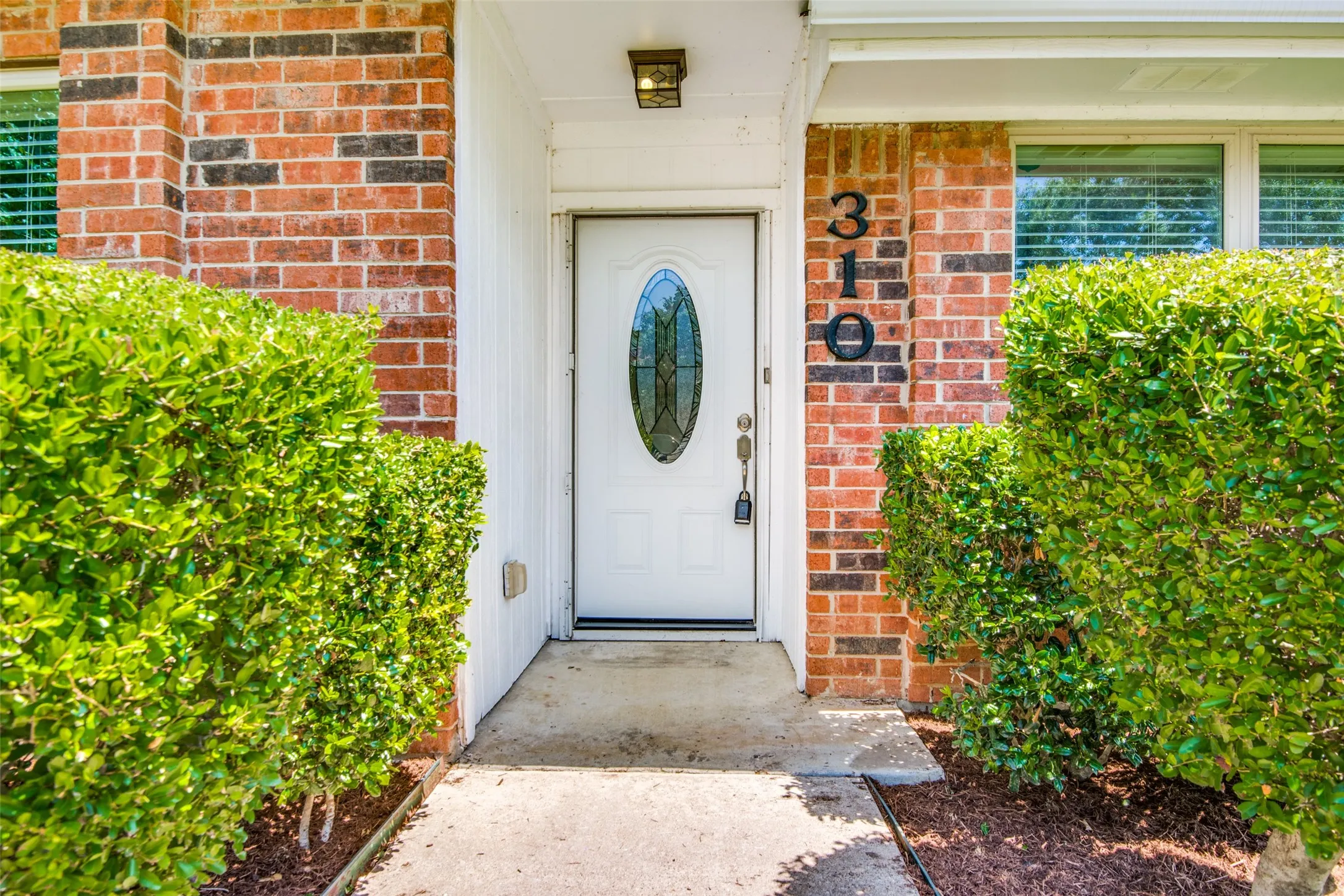 Brick exterior entry featuring a white door with an oval decorative glass insert, concrete walkway, and established shrubbery