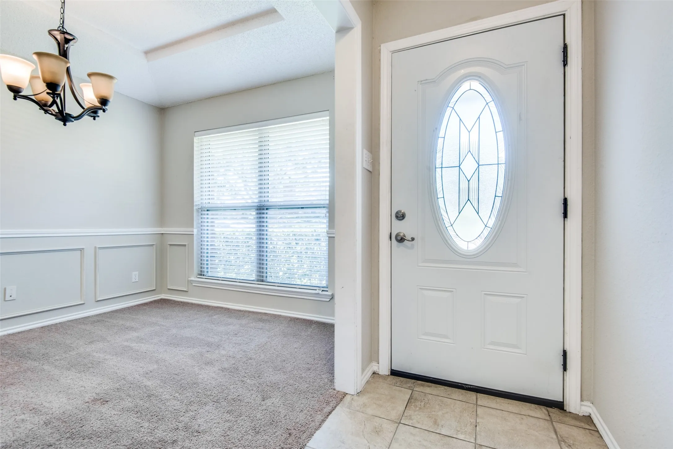 Entryway featuring a decorative oval glass front door, tile flooring, and a formal dining space with panel wainscoting, a chandelier, and carpet flooring