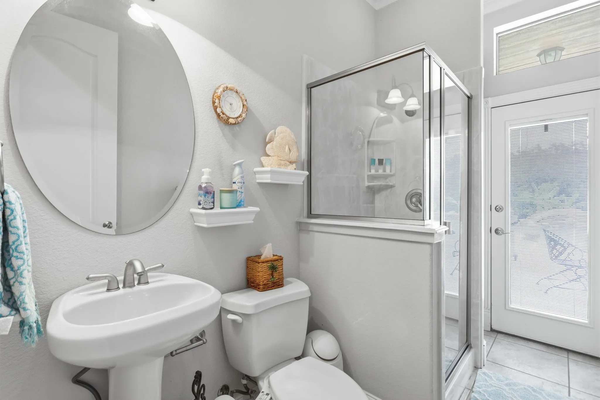 Pedestal sink with a brushed nickel faucet, large oval mirror, glass-enclosed shower with tiled walls, and a full-light exterior door