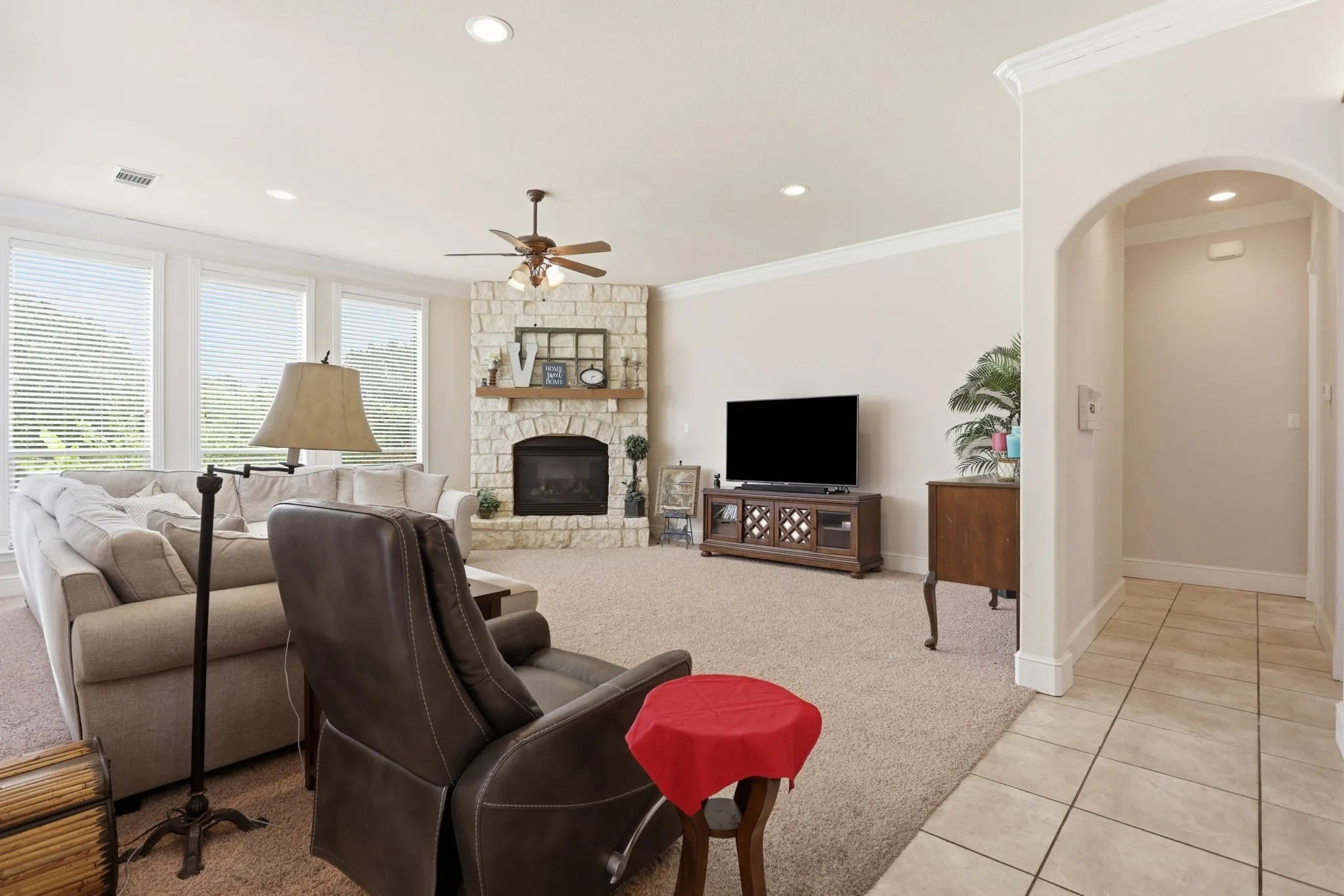 Spacious living area featuring a stone fireplace, crown molding, abundant natural light from multiple windows, recessed lighting, and an arched entryway