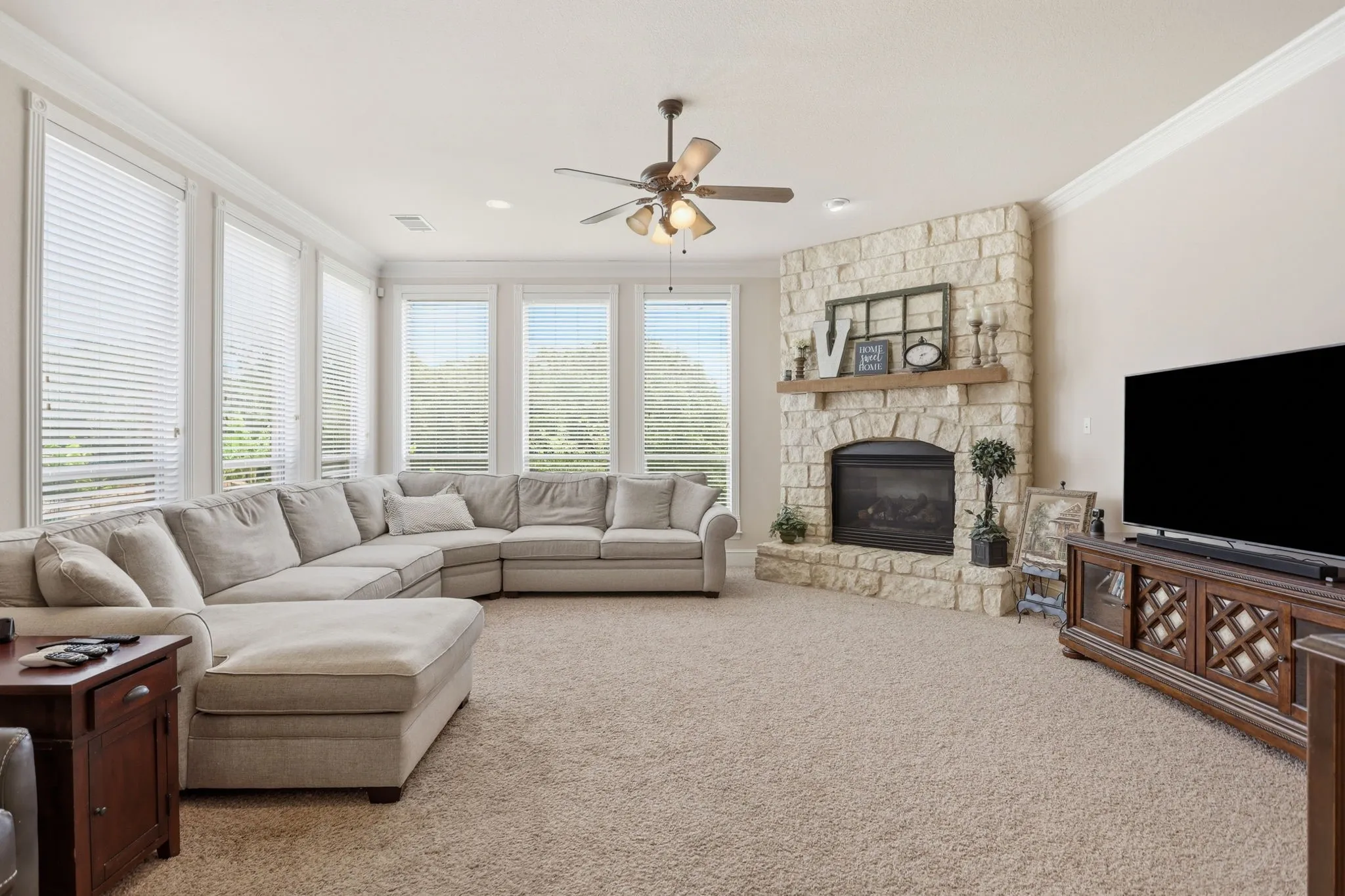 Living area featuring a stone fireplace with a wood mantel, light-colored carpeting, a ceiling fan, crown molding, and multiple windows with horizontal blinds
