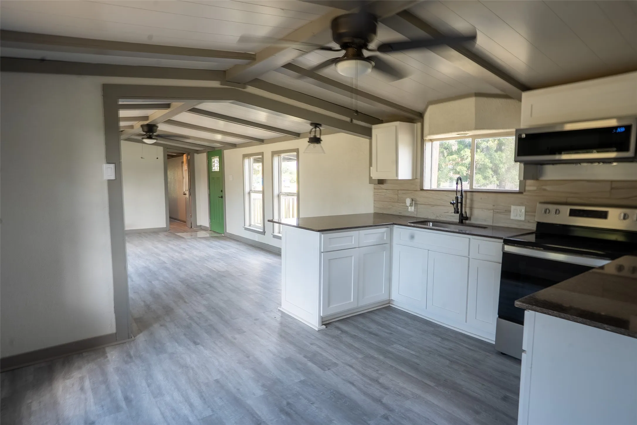 Open-concept kitchen and living area featuring wood-finish flooring, exposed ceiling beams, white cabinetry, dark countertops, and stainless steel appliances