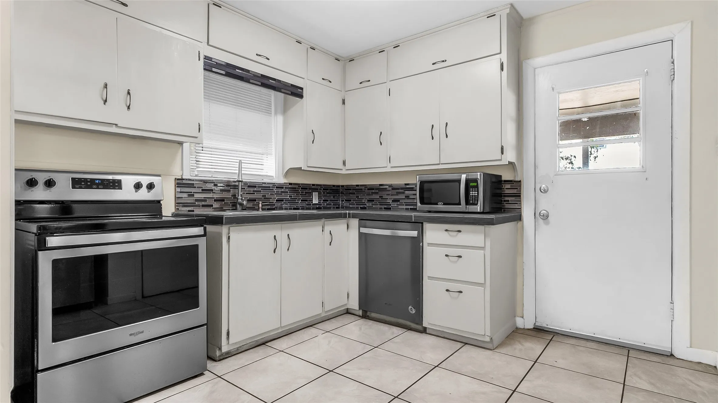Kitchen featuring white cabinetry, a mosaic tile backsplash, stainless steel appliances, dark countertops, and ceramic tile flooring