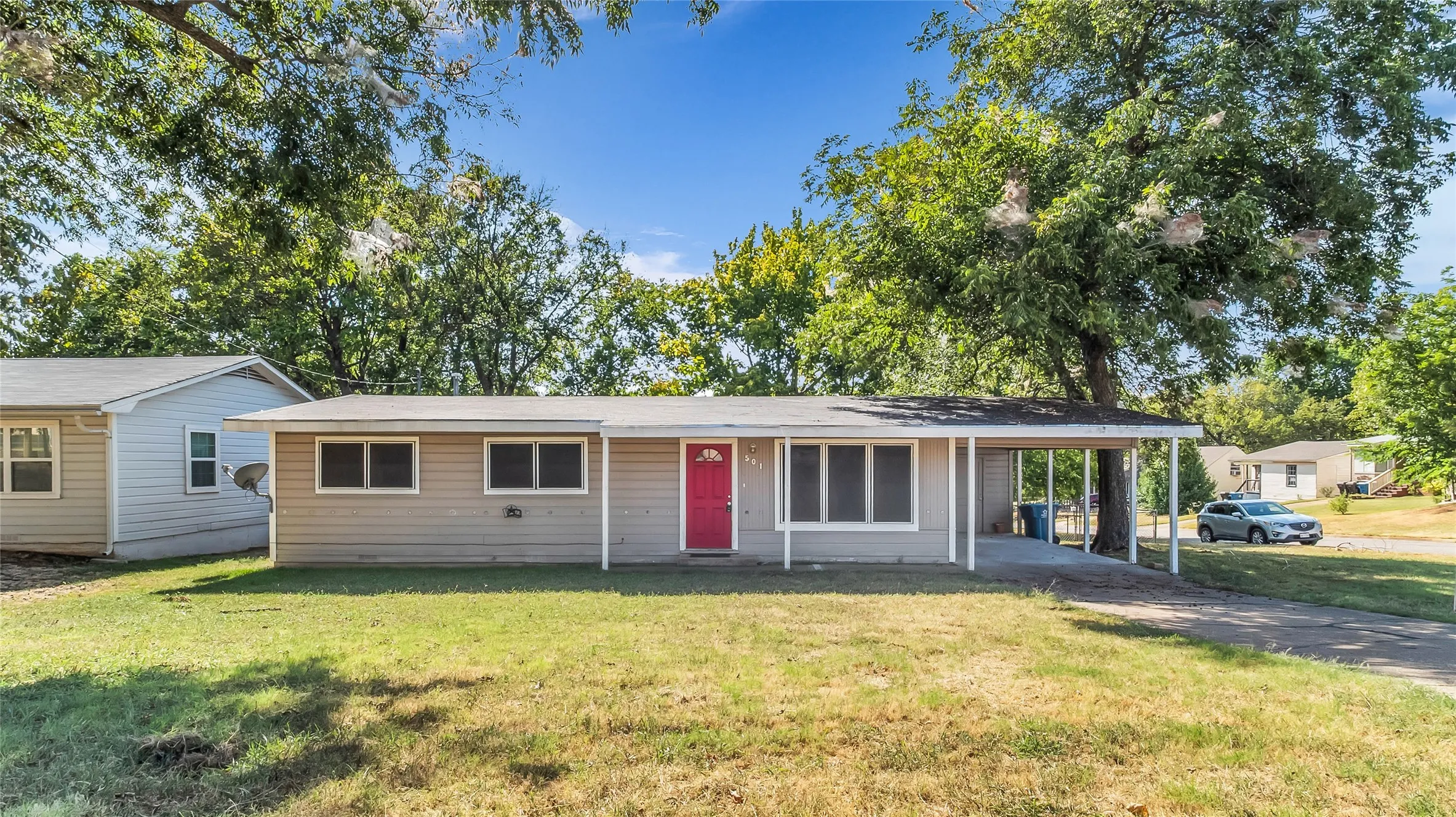Single-story residence featuring light-colored siding, a vibrant red entry door, a covered carport, and a paved driveway