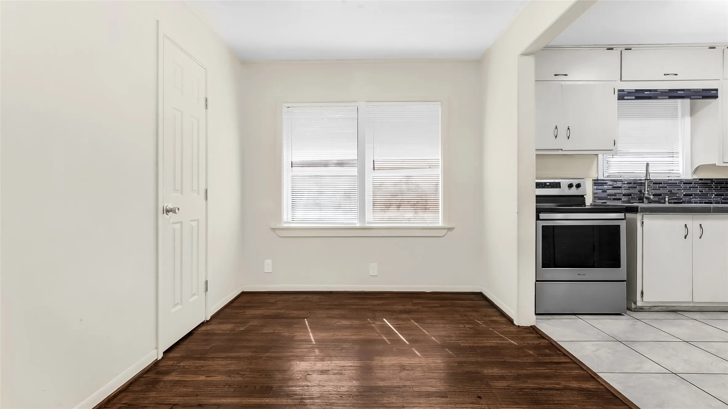 Rich wood-finish flooring, white paneled interior door, and a kitchen featuring white cabinetry, a tiled backsplash, and stainless steel appliances