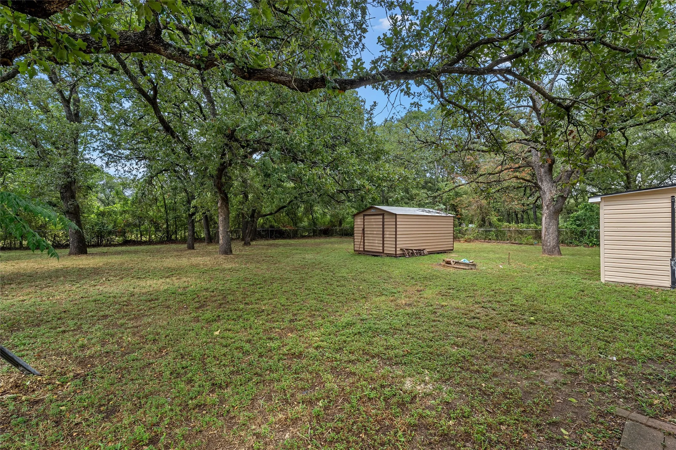 Expansive outdoor space featuring a large lawn area, mature trees providing natural canopy, and two storage sheds for utility