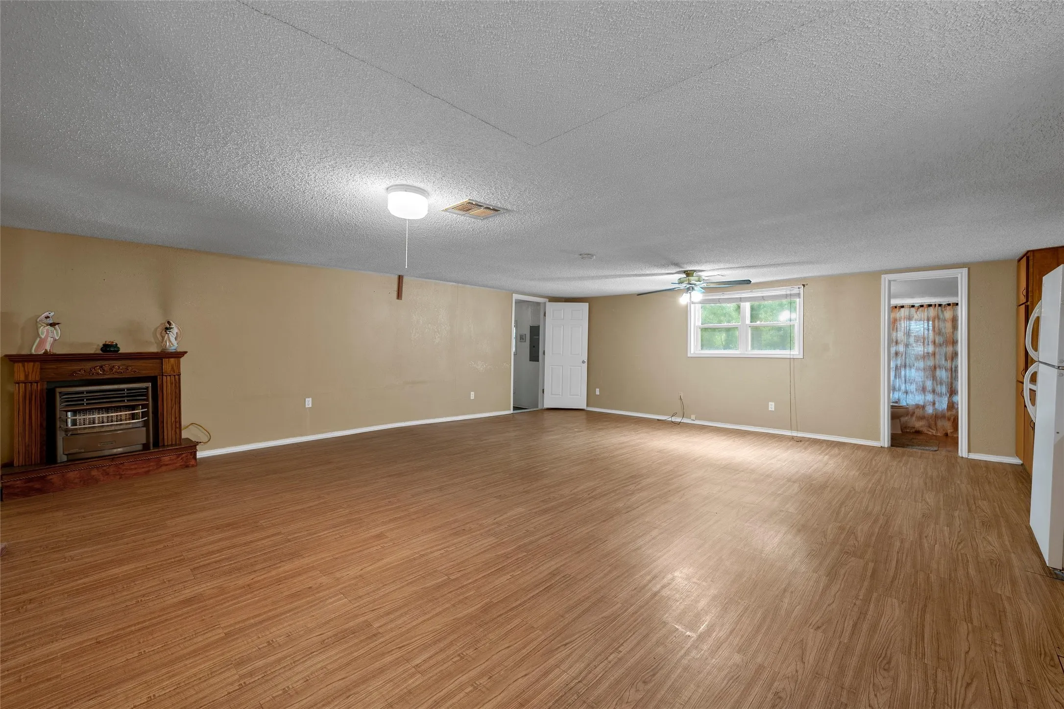 Spacious interior room featuring wood-finish flooring, a built-in fireplace with a decorative mantel, a ceiling fan, and ample natural light from the window