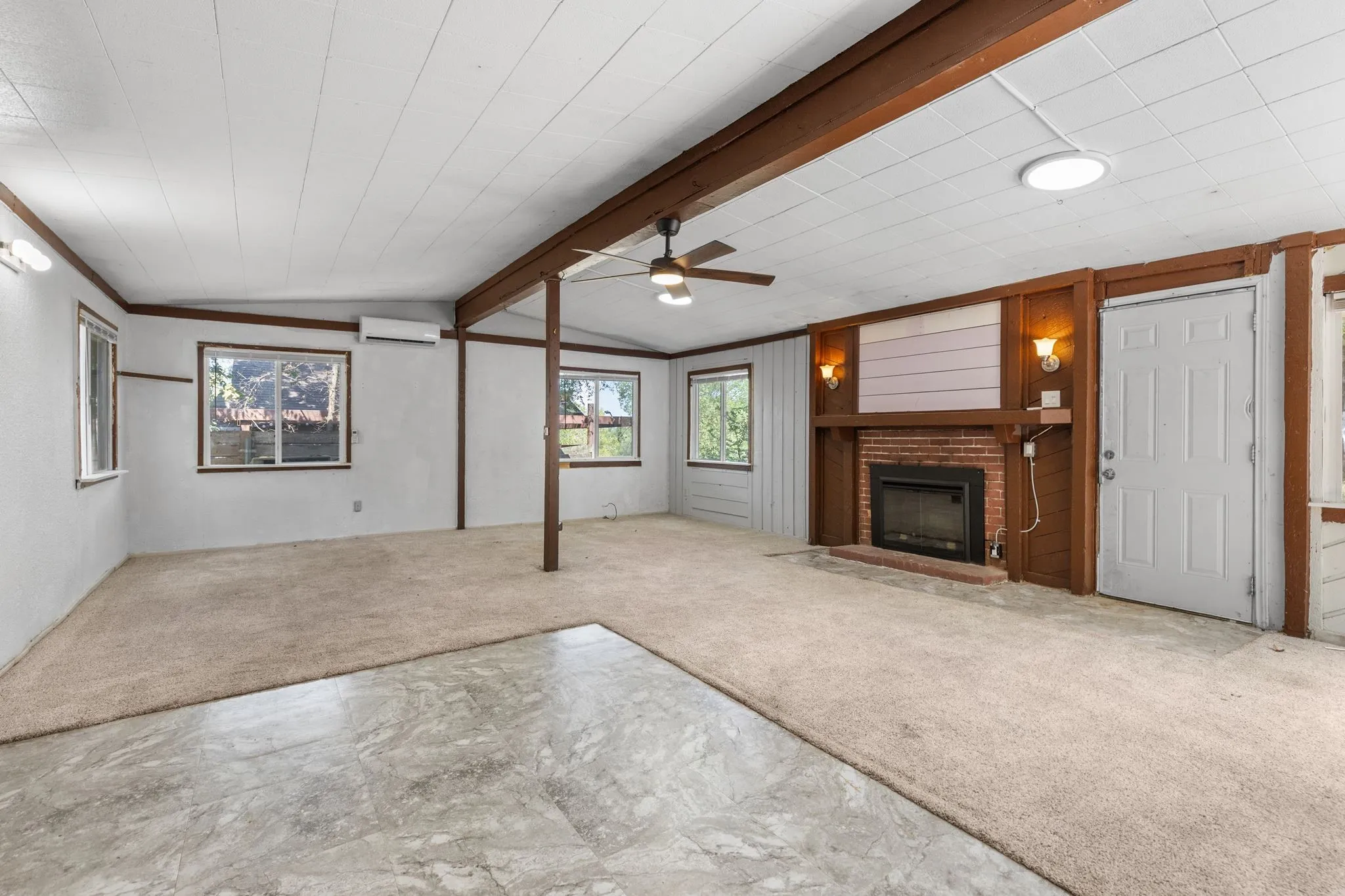 Brick fireplace with wood mantel, ceiling fan with integrated lighting, wood-finish beams, and carpeted flooring