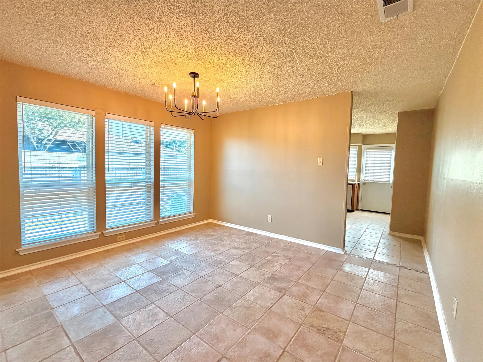 Spacious room featuring neutral-toned tile flooring, three large windows with blinds, a contemporary chandelier, and light-colored walls