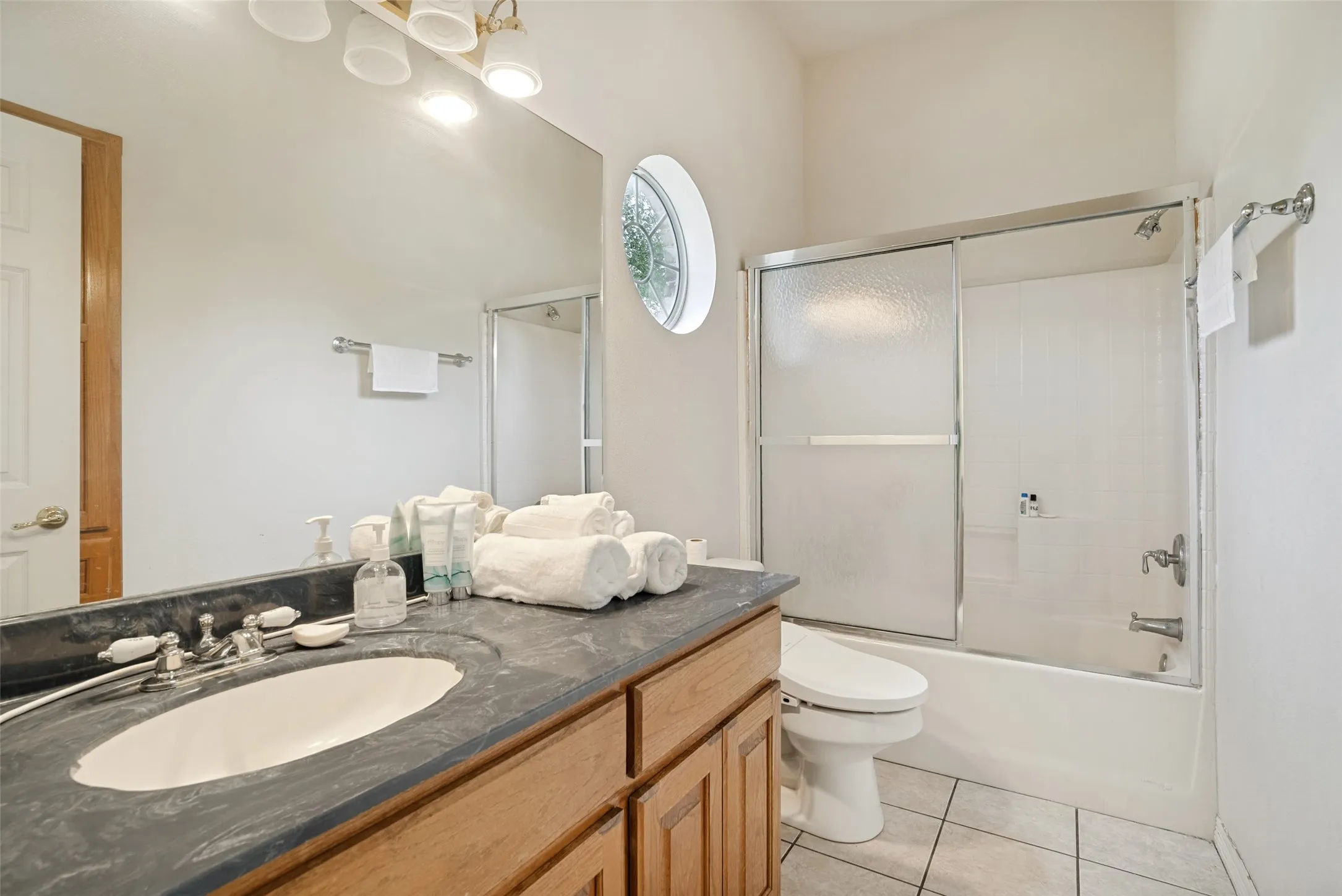 Full bathroom with wood cabinetry and a dark countertop, an oval-shaped sink, a tub-shower combination with sliding frosted glass doors, and light-toned floor tiling