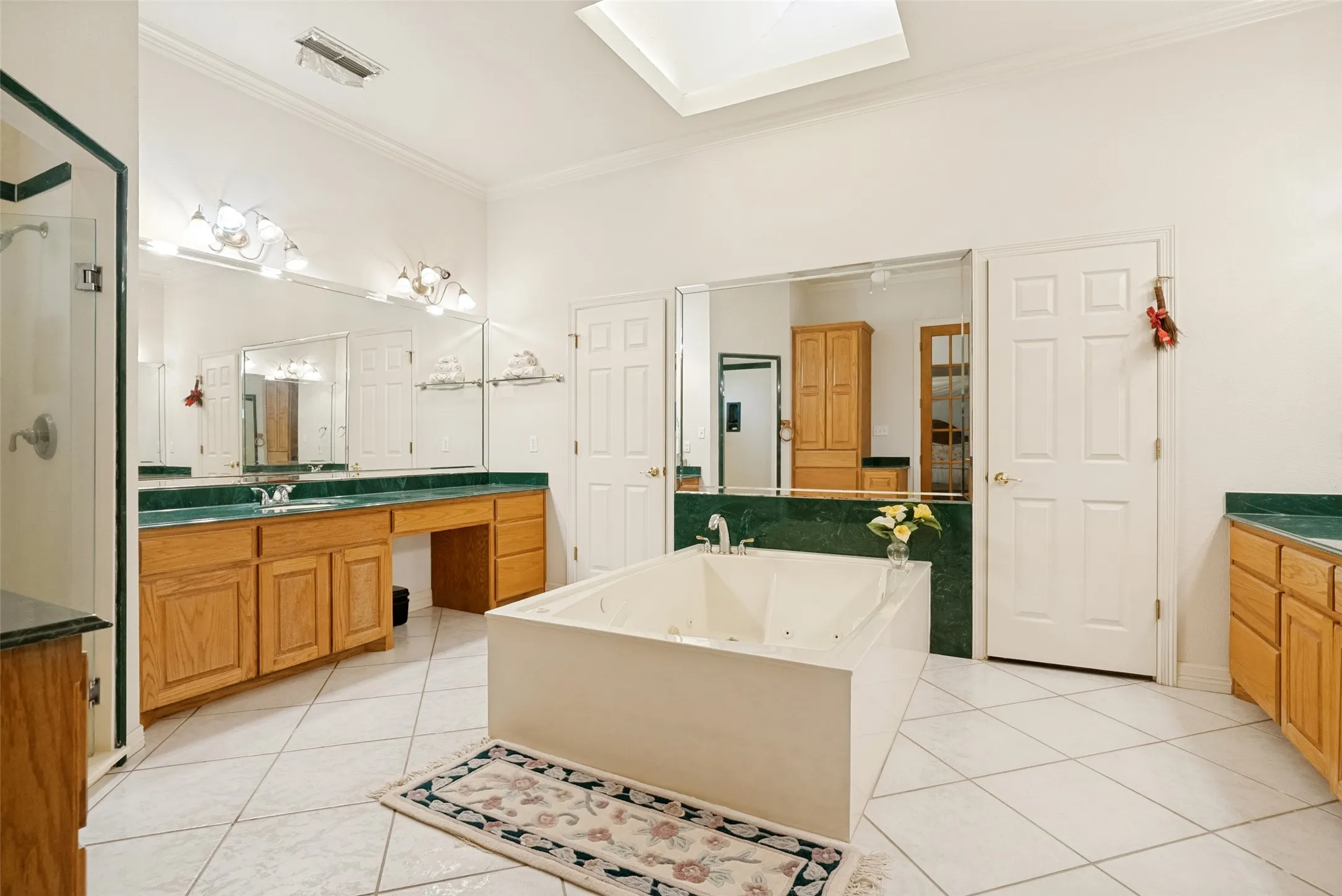 Spacious bathroom featuring a large skylight, built-in jetted soaking tub, dual vanities with integrated sinks, wood-finish cabinetry, and a glass-enclosed shower